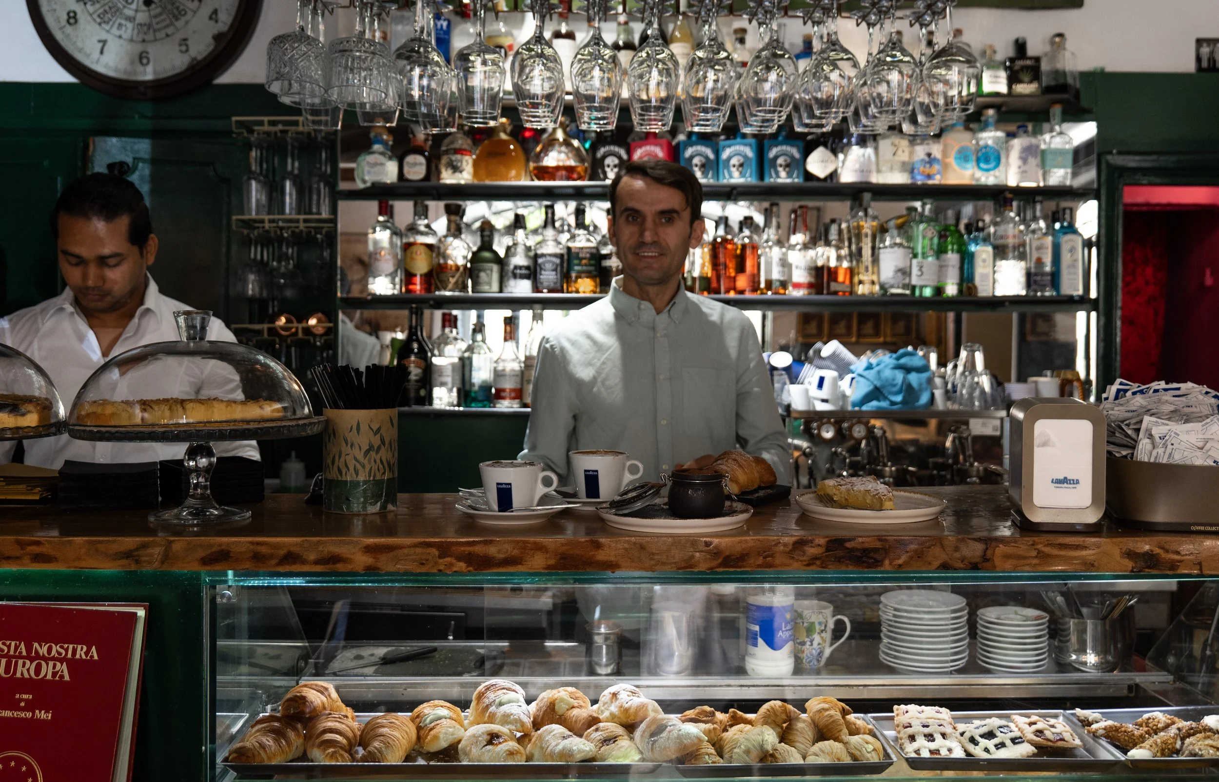Barista dietro il bancone di un bar con pasticcini e cioccolata calda, scaffali con bottiglie di alcolici e bicchieri appesi sopra.