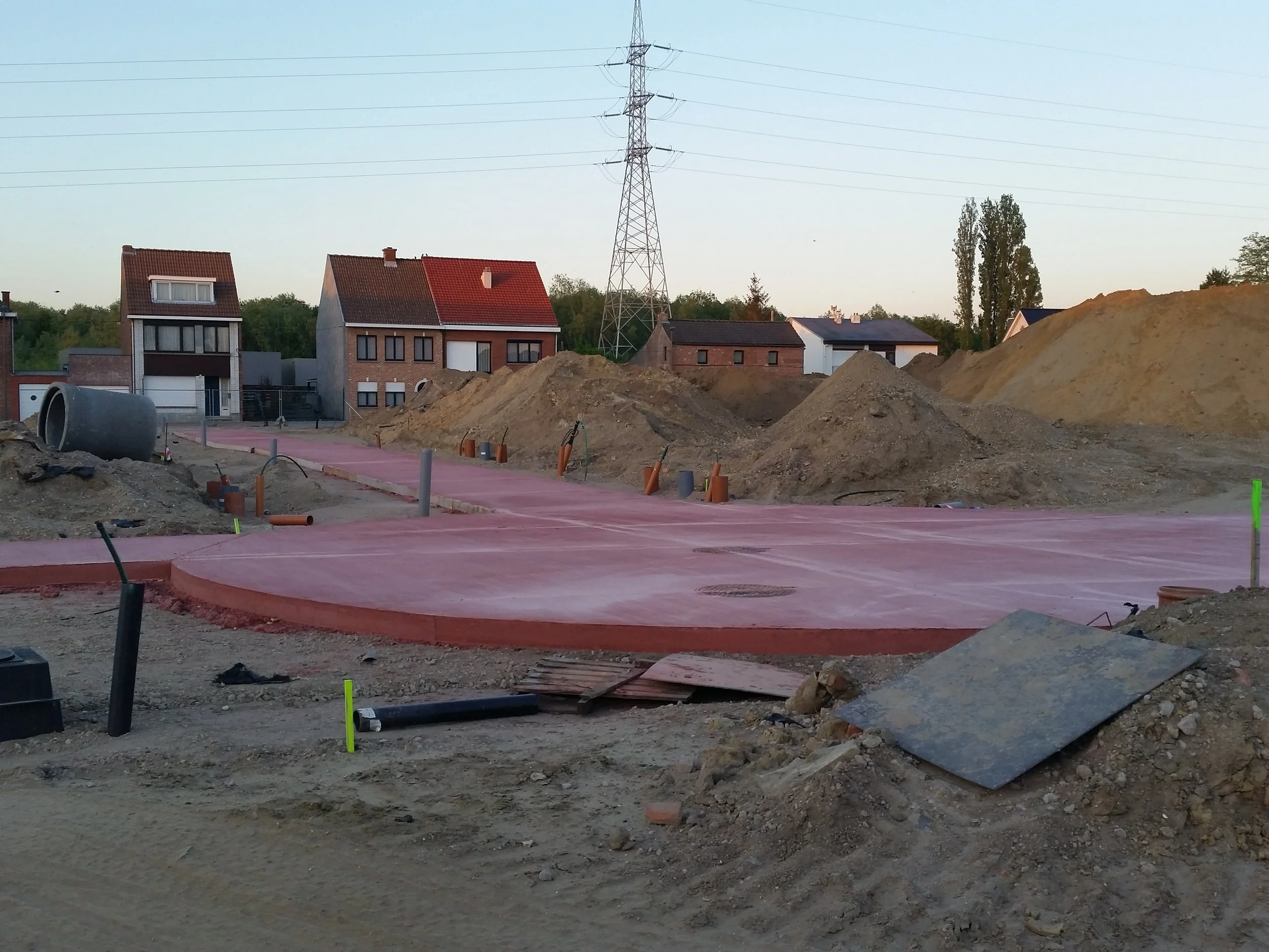 Bouwterrein met afgewerkte en nog te voltooien delen, bouwmaterialen, grondhopen, en huizen op de achtergrond.