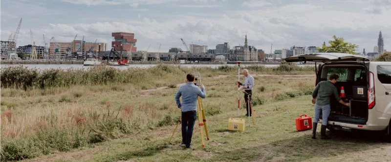 Mannen voeren een landmeting uit op een grasveld met een stad op de achtergrond, terwijl een andere man enkele meetapparaten en koffers bij een geparkeerde auto plaatst.