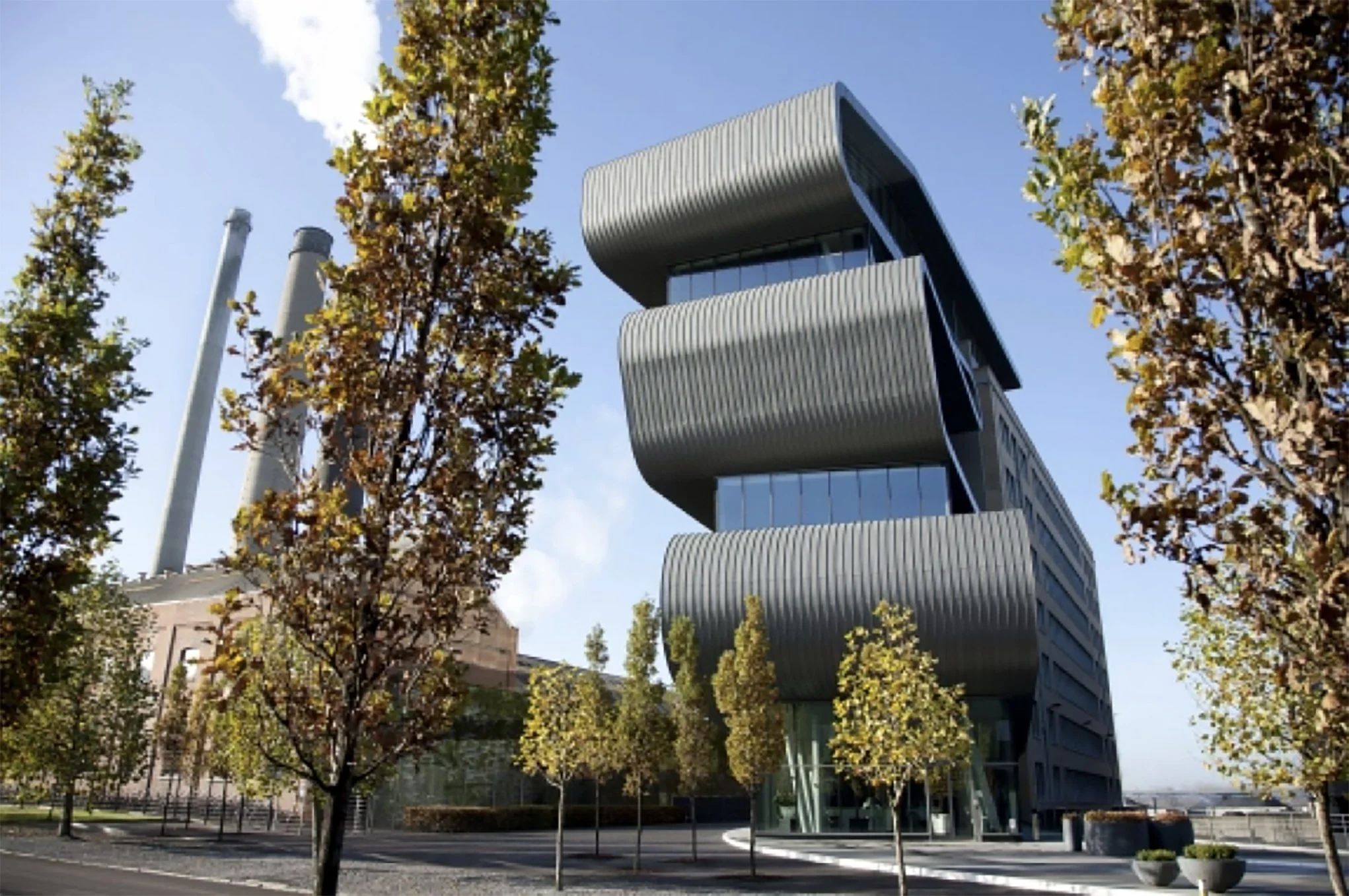 Moderne gebouw met afgeronde, horizontale gevelbekleding, omringd door bomen met herfstkleuren, onder een blauwe lucht.