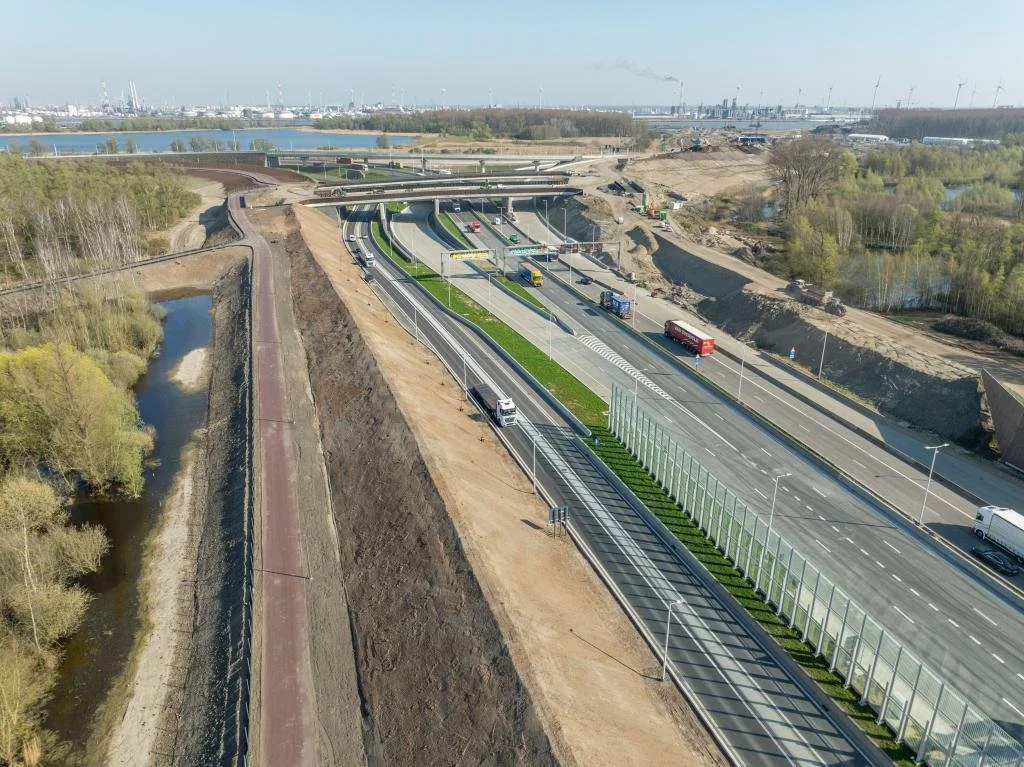 Luchtfoto van een snelweg en fietsroute onder constructie, met voertuigen en een groenvoorziening in de middenberm, omringd door natuur en water.