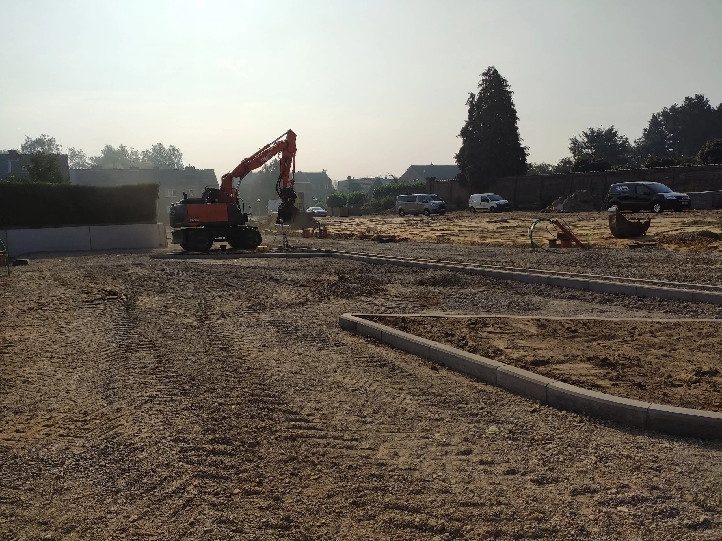Bouwplaats met een graafmachine, een asfaltdak en enkele geparkeerde auto's op de achtergrond.