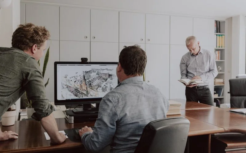 Drie mannen bespreken plannen in een vergaderruimte, één kijkt op een computerscherm met een bouwproject en een andere leest een boek.