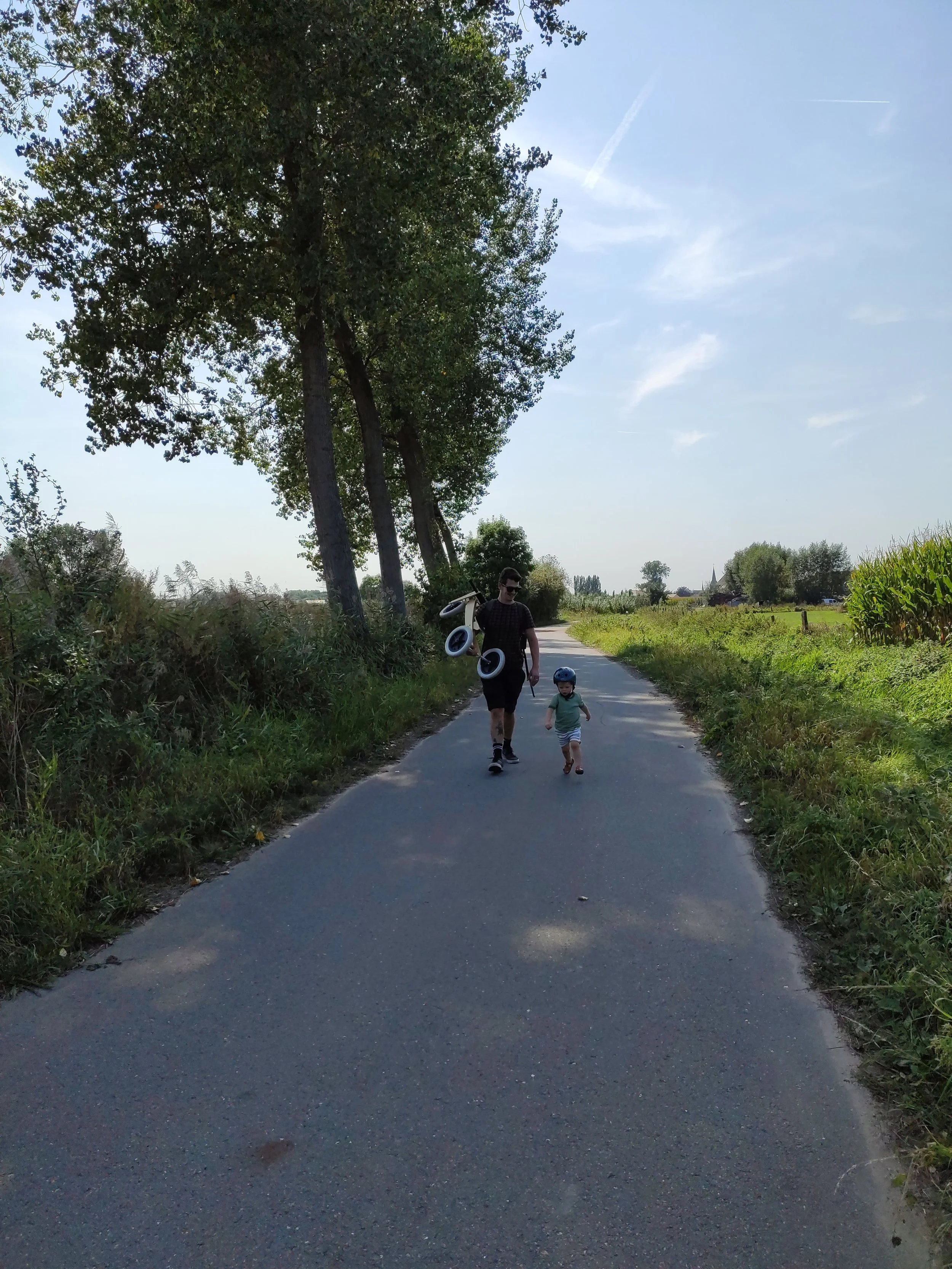 Een man en een kind lopen samen op een smal asfaltweggetje in een landelijke omgeving. De man draagt een skateboard over zijn schouder en het kind draagt een helm.
