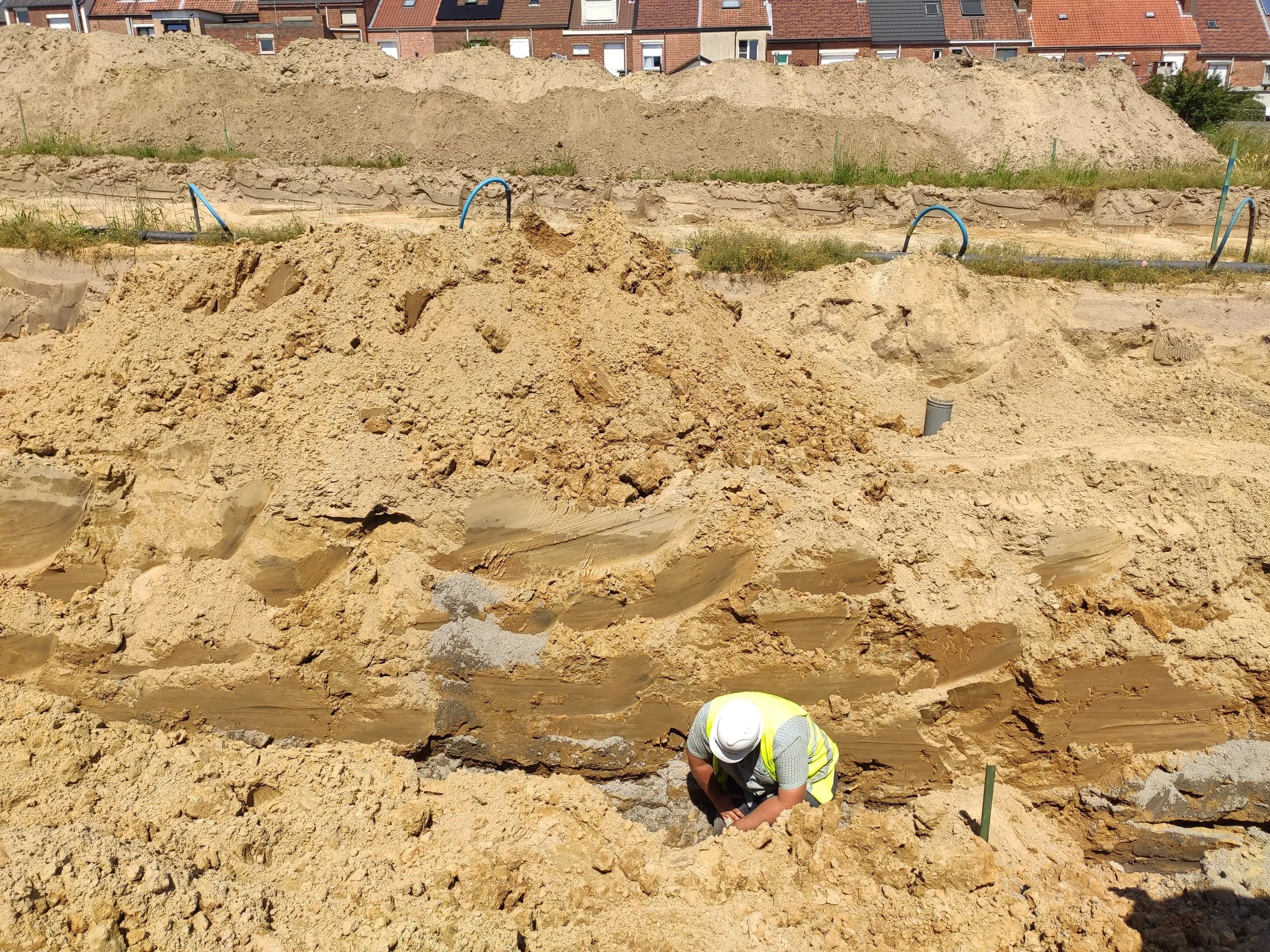Werknemer in geel veiligheidsvest en witte helm onderzoekt de ondergrond van een bouwplaats met zand en zandafzetting, nabij enkele buizen en in een stedelijke omgeving.