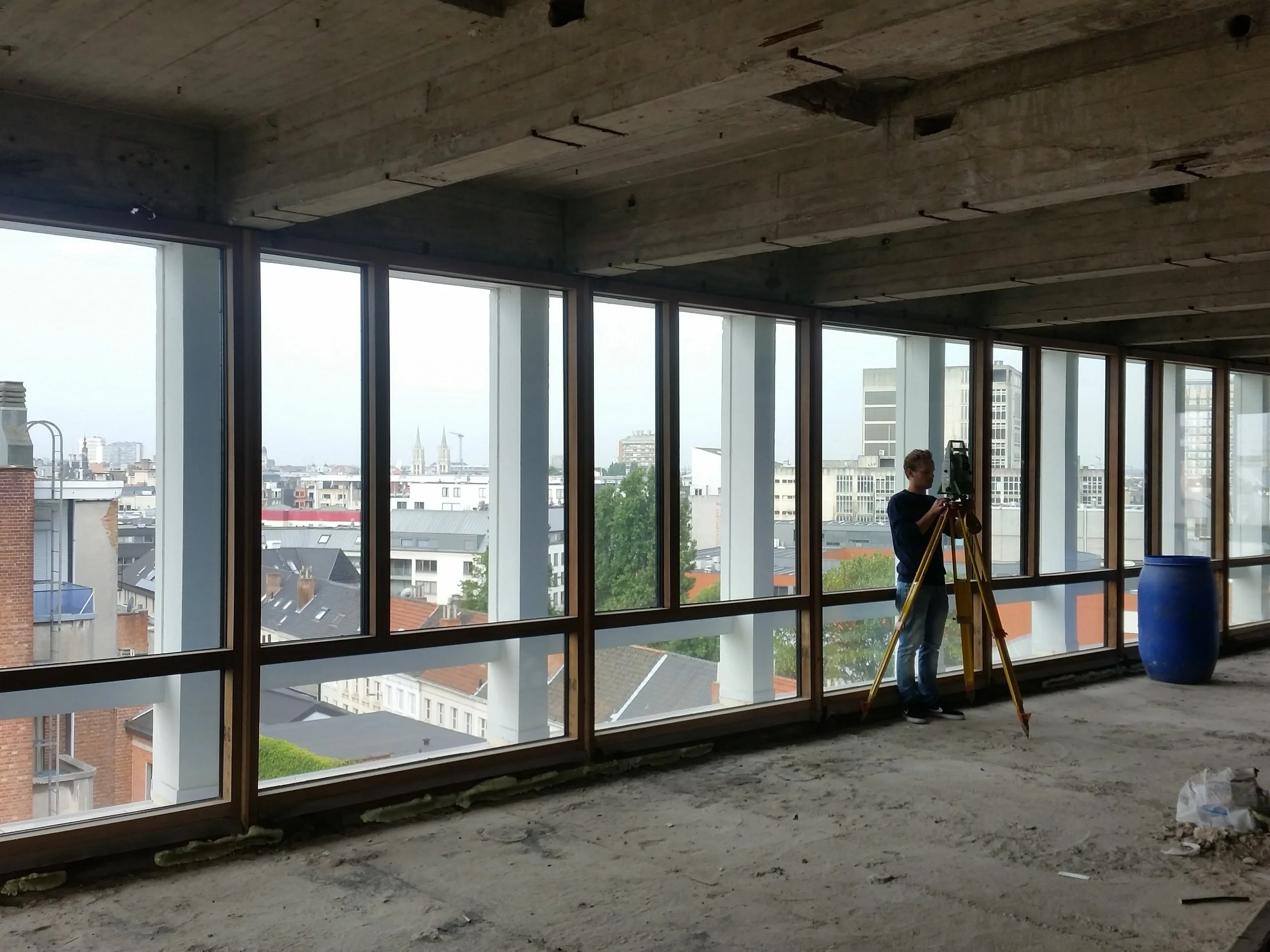 Binnen een onvoltooide ruimte met grote ramen, een man gebruikt een instrument op een statief, mogelijk voor bouwwerkzaamheden of bouwinspectie.