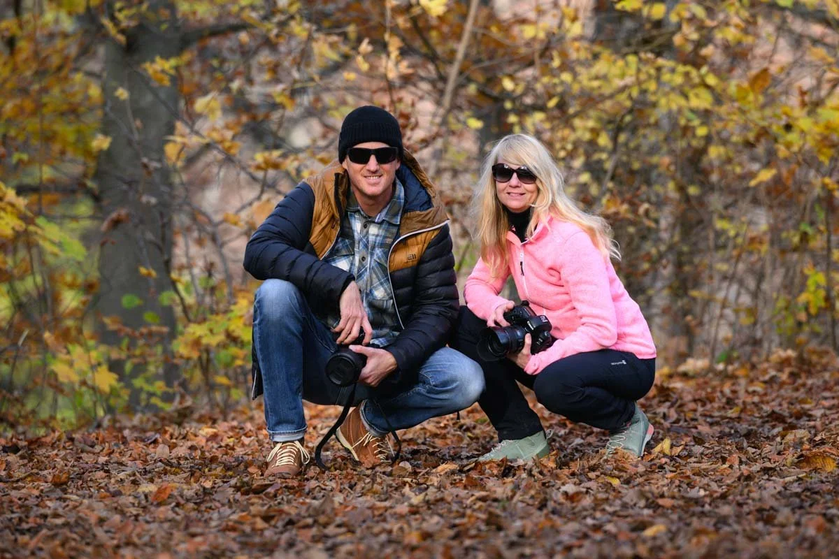 Frédéric Diserens und Isabelle Egli knien in einem herbstlichen Wald, beide halten Kameras und sind mit Sonnenbrillen bekleidet.