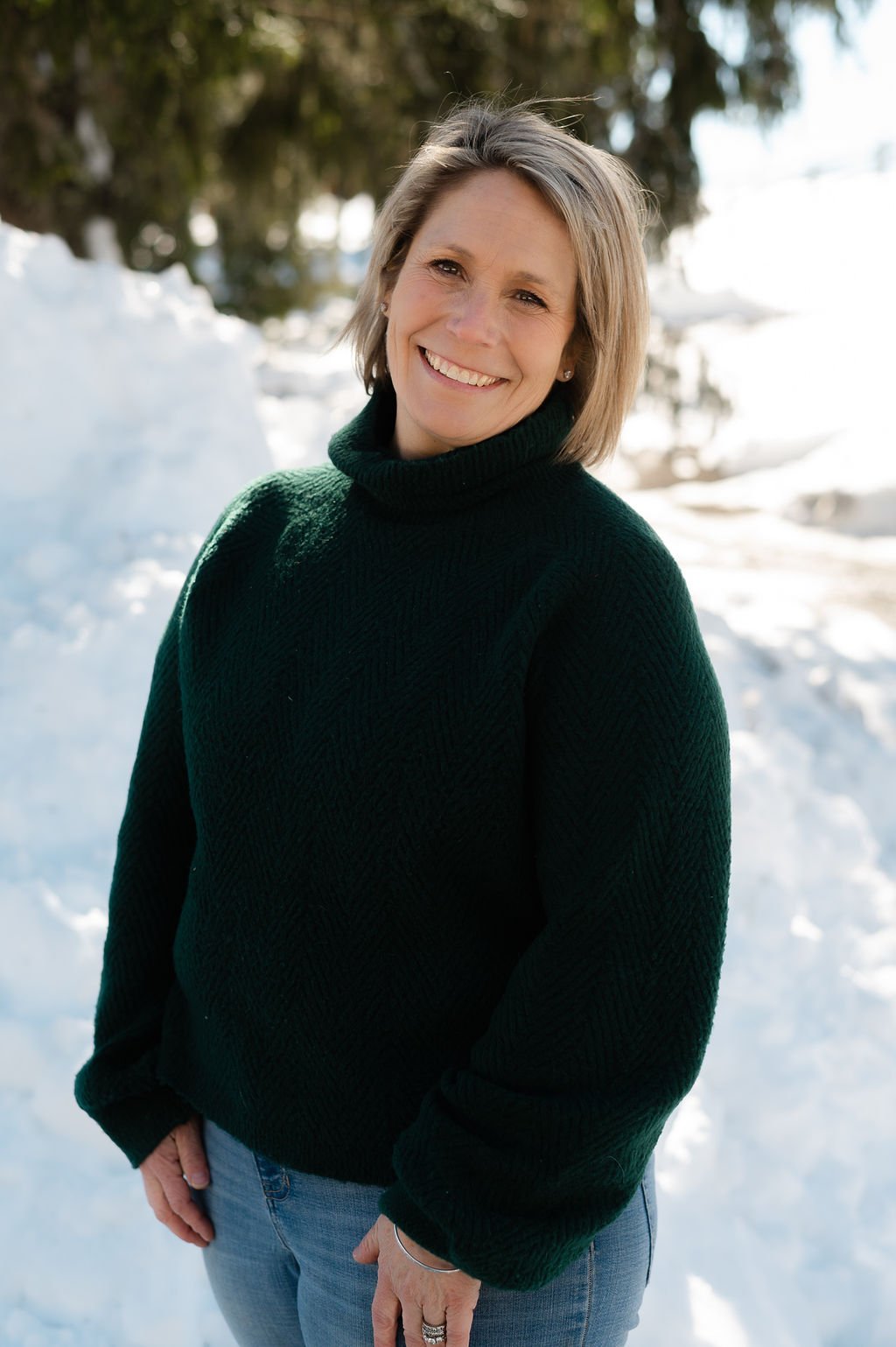 A woman with short blonde hair and a big smile, wearing a dark green turtleneck sweater and blue jeans, standing outdoors in a snowy landscape with trees in the background.