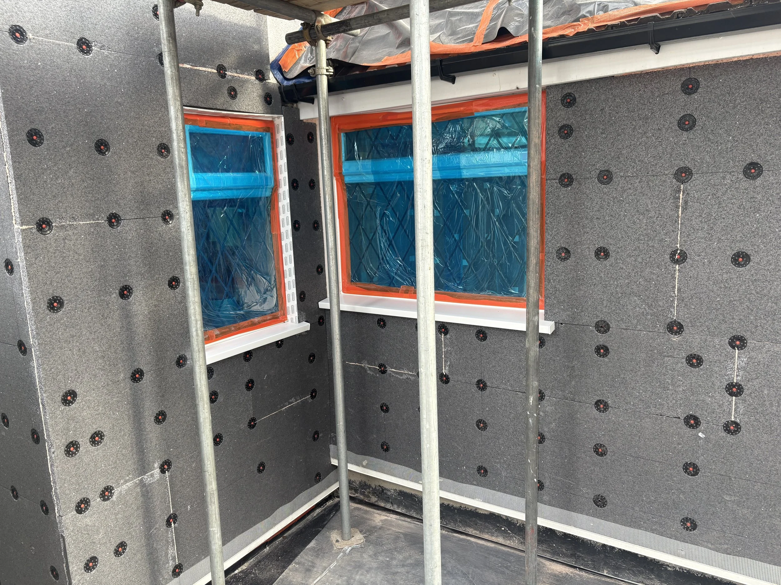 Interior of a building under construction with black insulation boards, two small windows covered with plastic, a steel scaffold, and orange sealing around the windows.