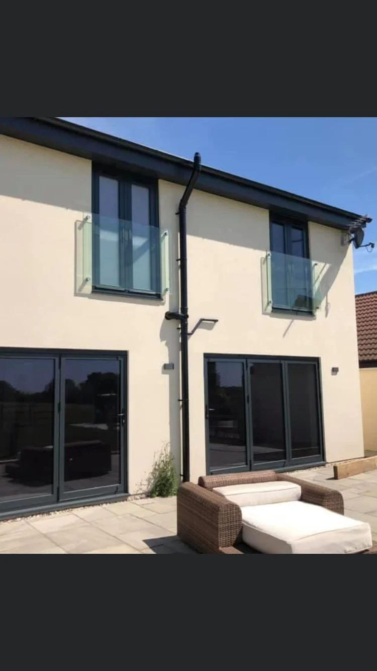 Modern two-story house with beige exterior, large glass sliding doors, and windows. Balcony with glass railing on the upper floor. Outdoor patio with wicker sofa and cushions, and plants nearby.