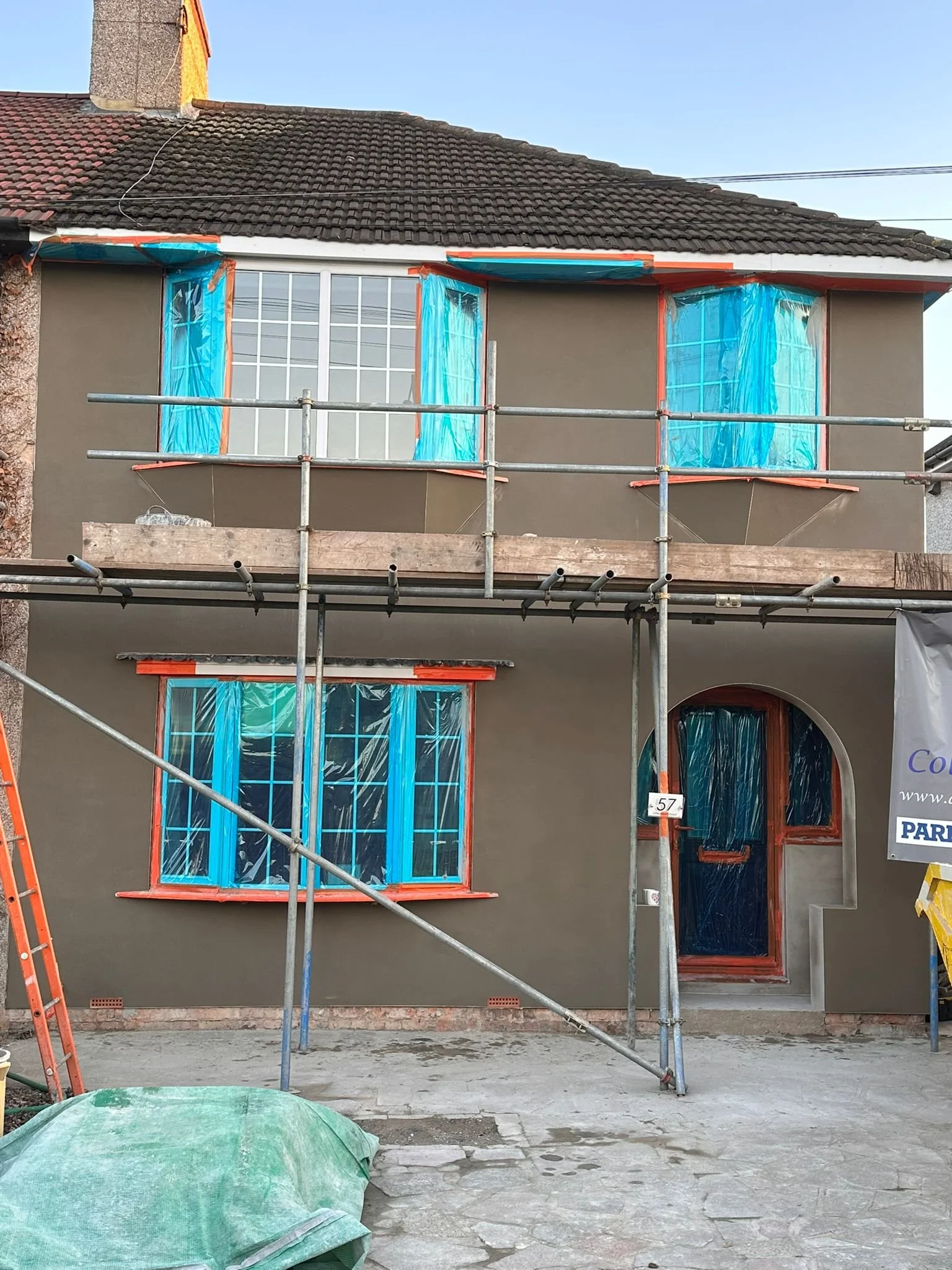 Exterior of a house with scaffolding in front, showing part of the roof, a window, and the sky.