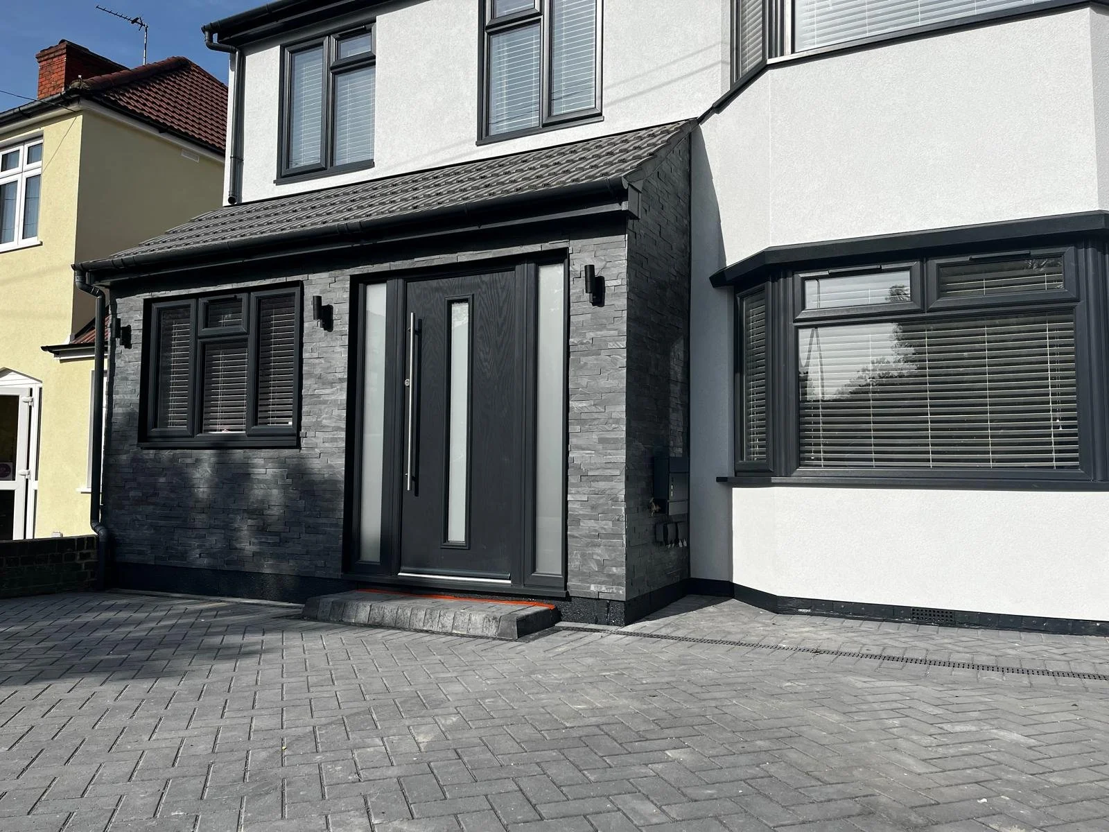 Modern house with black and white exterior, black front door with glass panels, black window shutters, bay window, brick-paved driveway, and black roof gutters