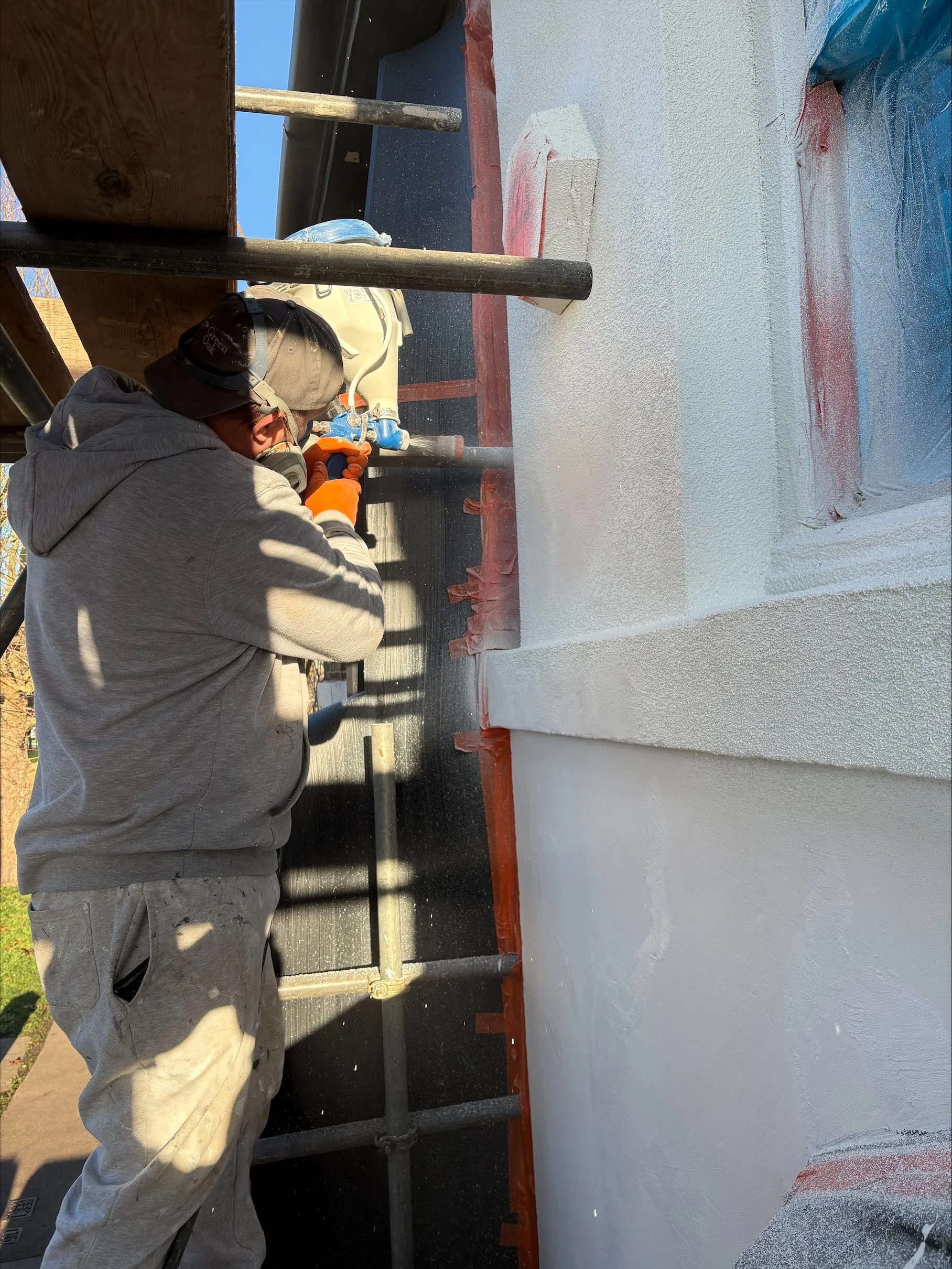 A person spray painting a building's exterior wall white, wearing a hoodie, safety goggles, and gloves, with scaffolding around them.