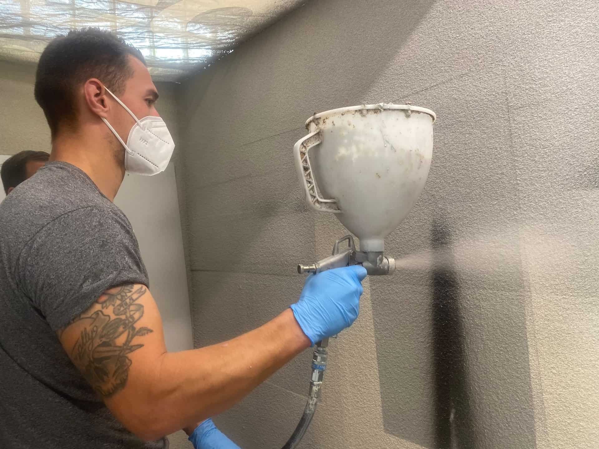 A worker covers a wall with CorkSol using a sprayer, wearing a mask