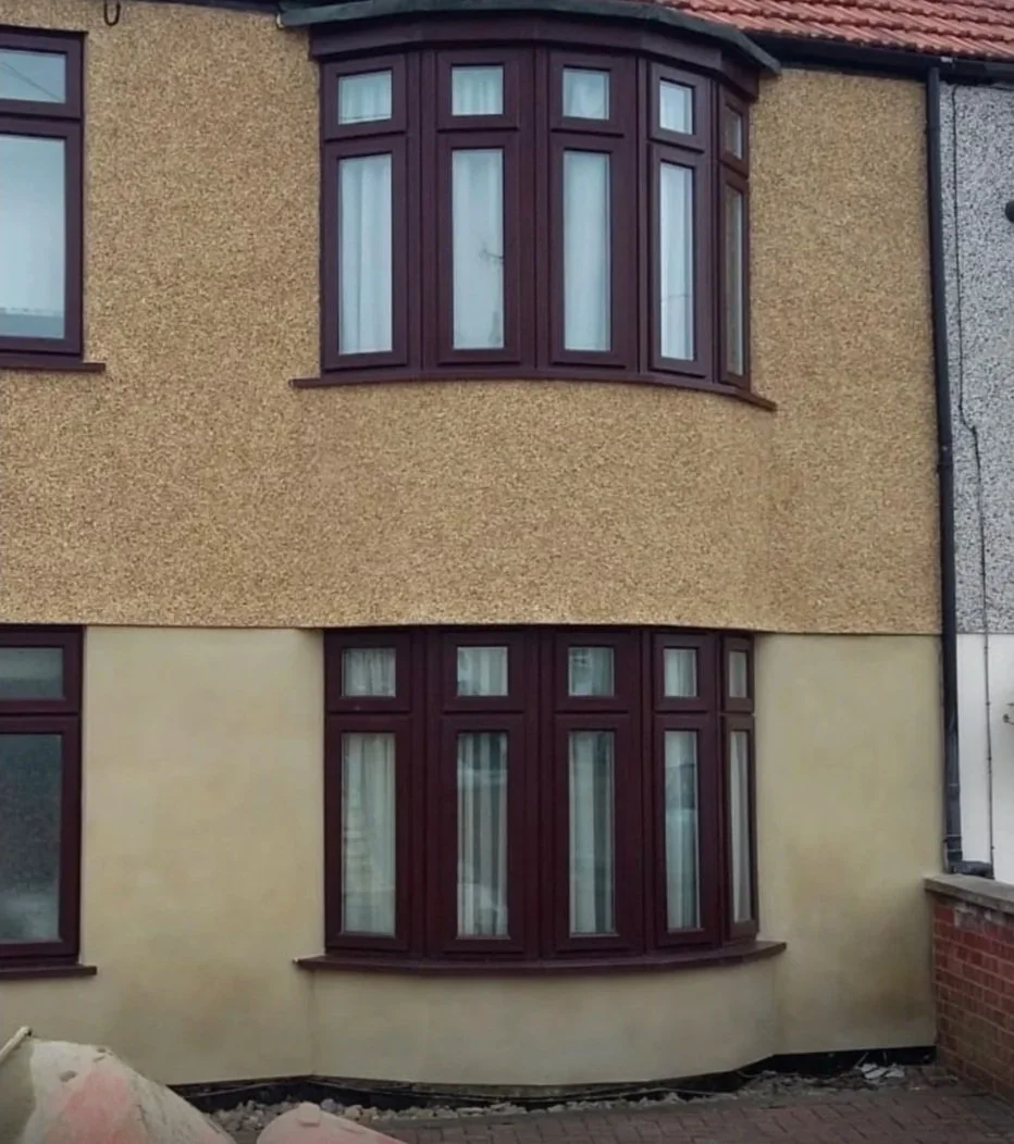 A two-story house with textured wall surfaces, featuring a large bay window on the upper floor and a smaller bay window on the ground floor, both framed with dark wood.