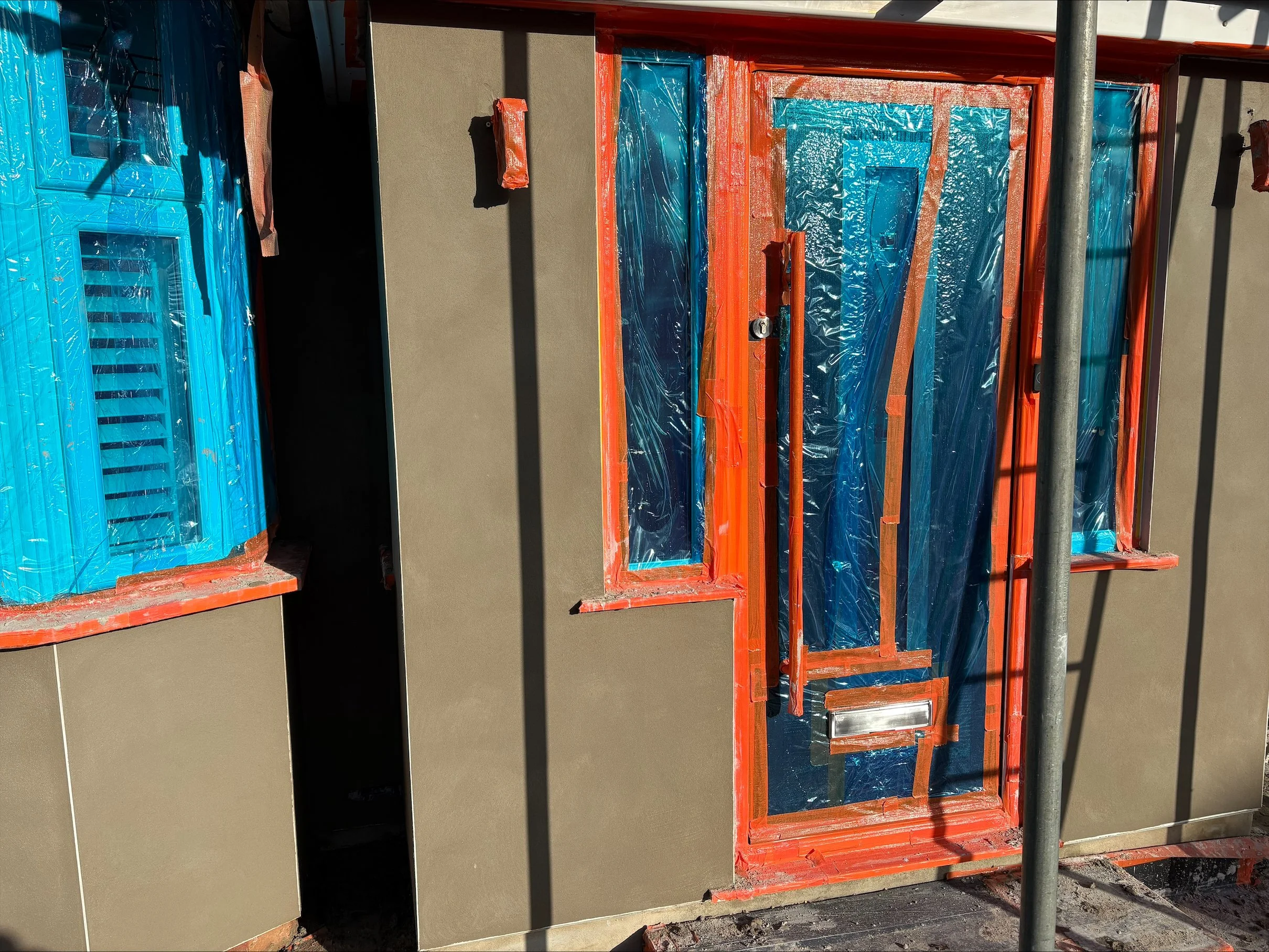 Building exterior under construction with beige walls, blue plastic-wrapped windows, and a front door with protective plastic and orange framing, surrounded by construction materials and scaffolding.