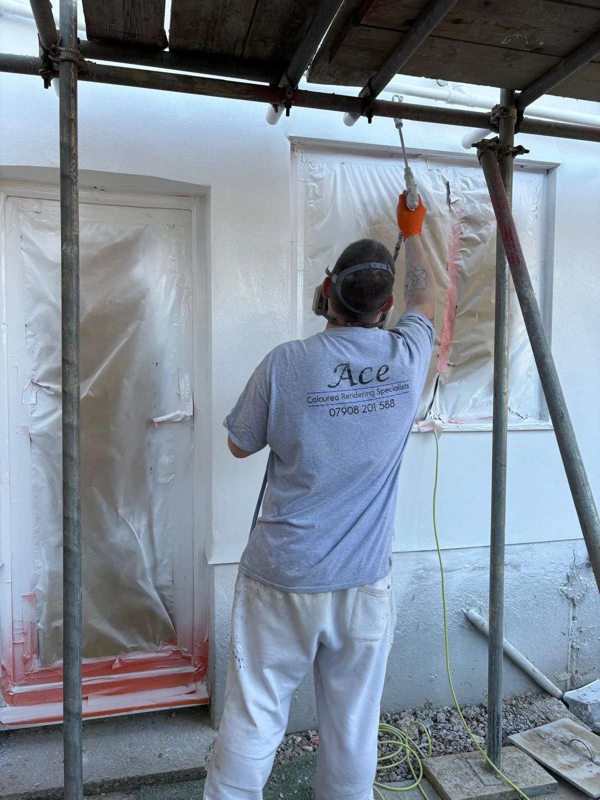 A construction worker painting or sealing around a window on a house exterior during renovation, wearing a gray t-shirt and white pants, with scaffolding around him.