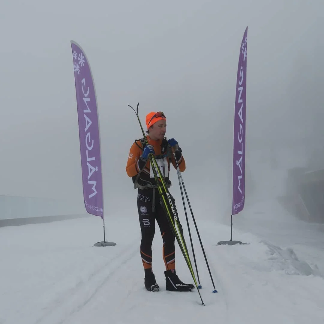 A person in winter sports gear standing on snow, holding ski poles, with two purple flags on either side. The person is dressed in black and orange with a backpack, helmet, and goggles.