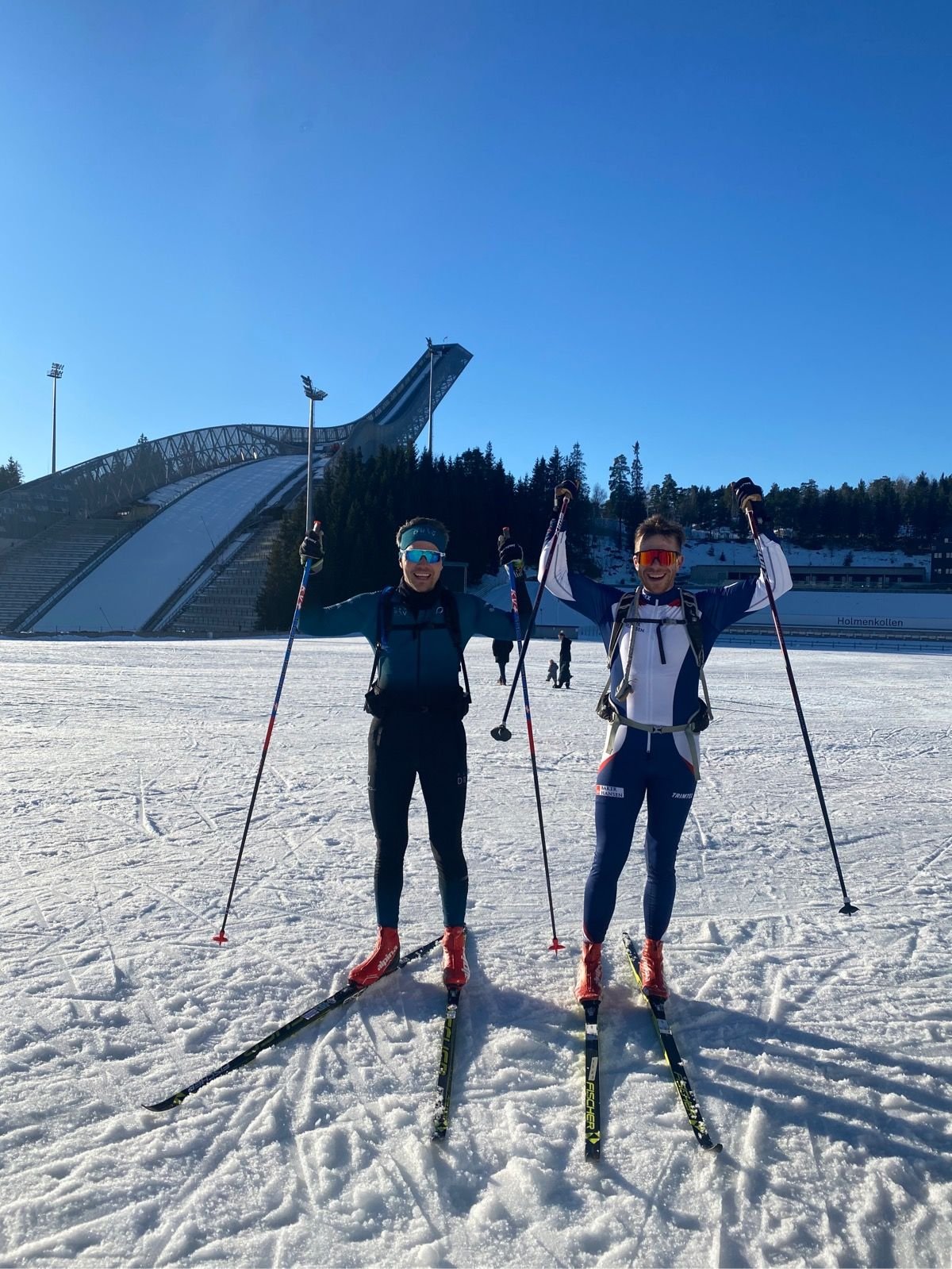 Two men in ski gear standing on snow with their arms raised, holding ski poles, smiling, in front of an Olympic ski jump stadium under a clear blue sky.