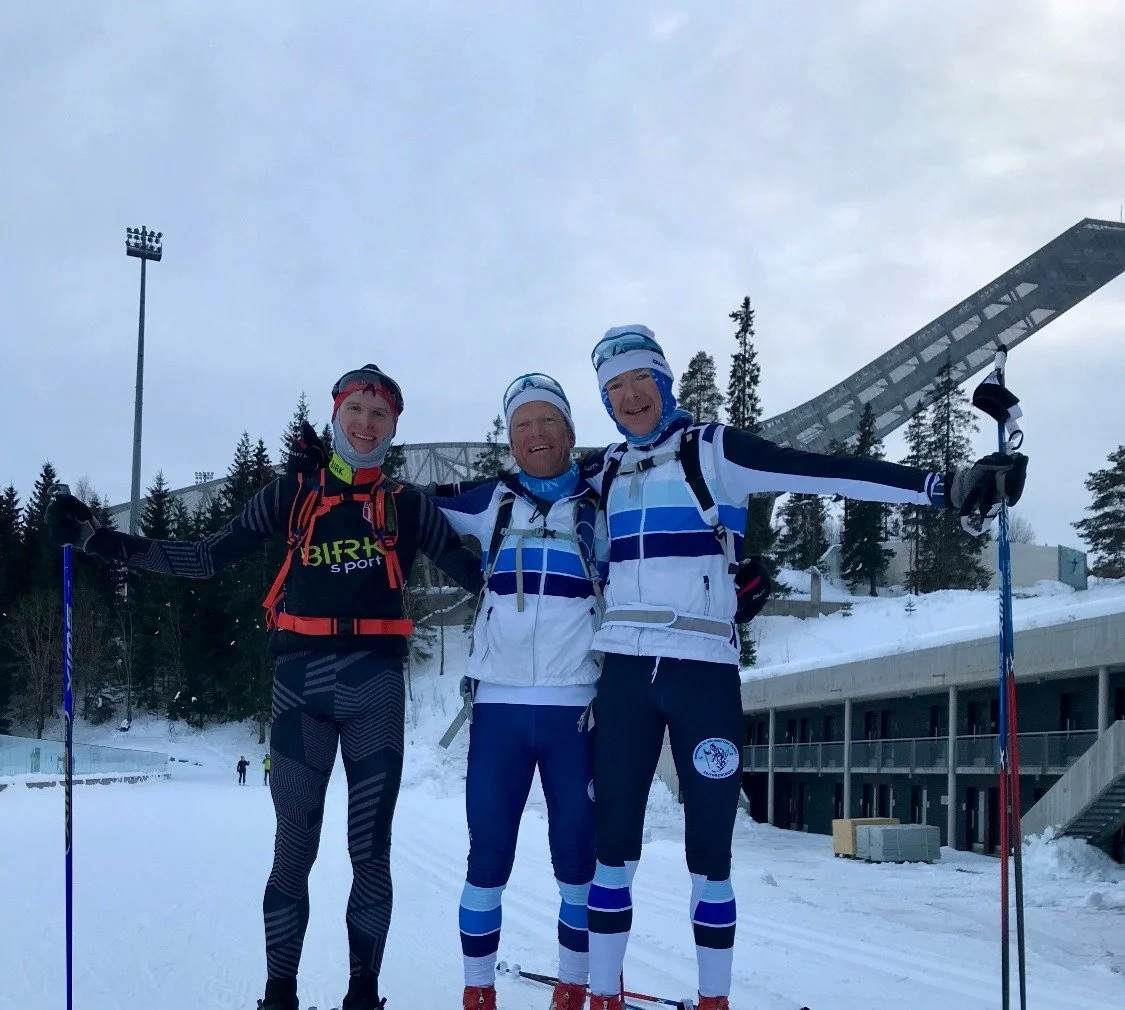 Three skiers standing together outside on snow, smiling with arms around each other, wearing snow gear and holding ski poles, with snow-covered trees and a ski jump in the background.