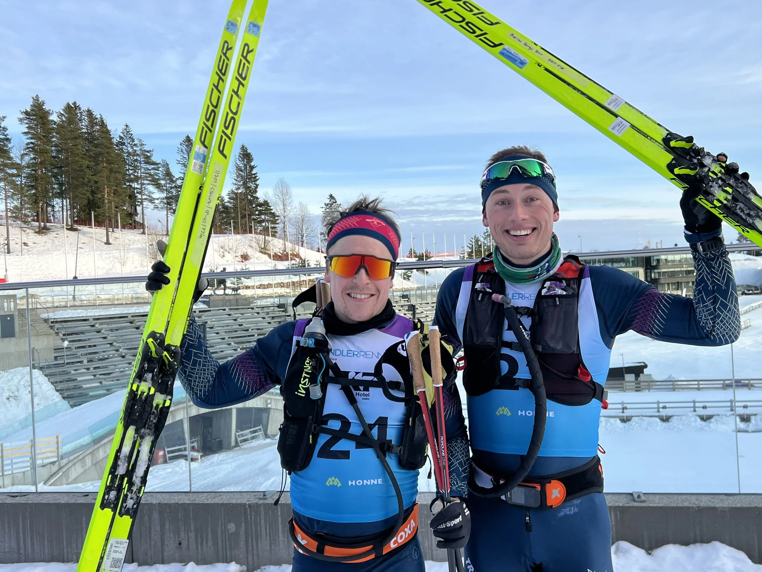 Two male ski mountaineers in race gear smiling and holding skis above their heads, standing in a snowy outdoor area with a ski stadium and trees in the background.