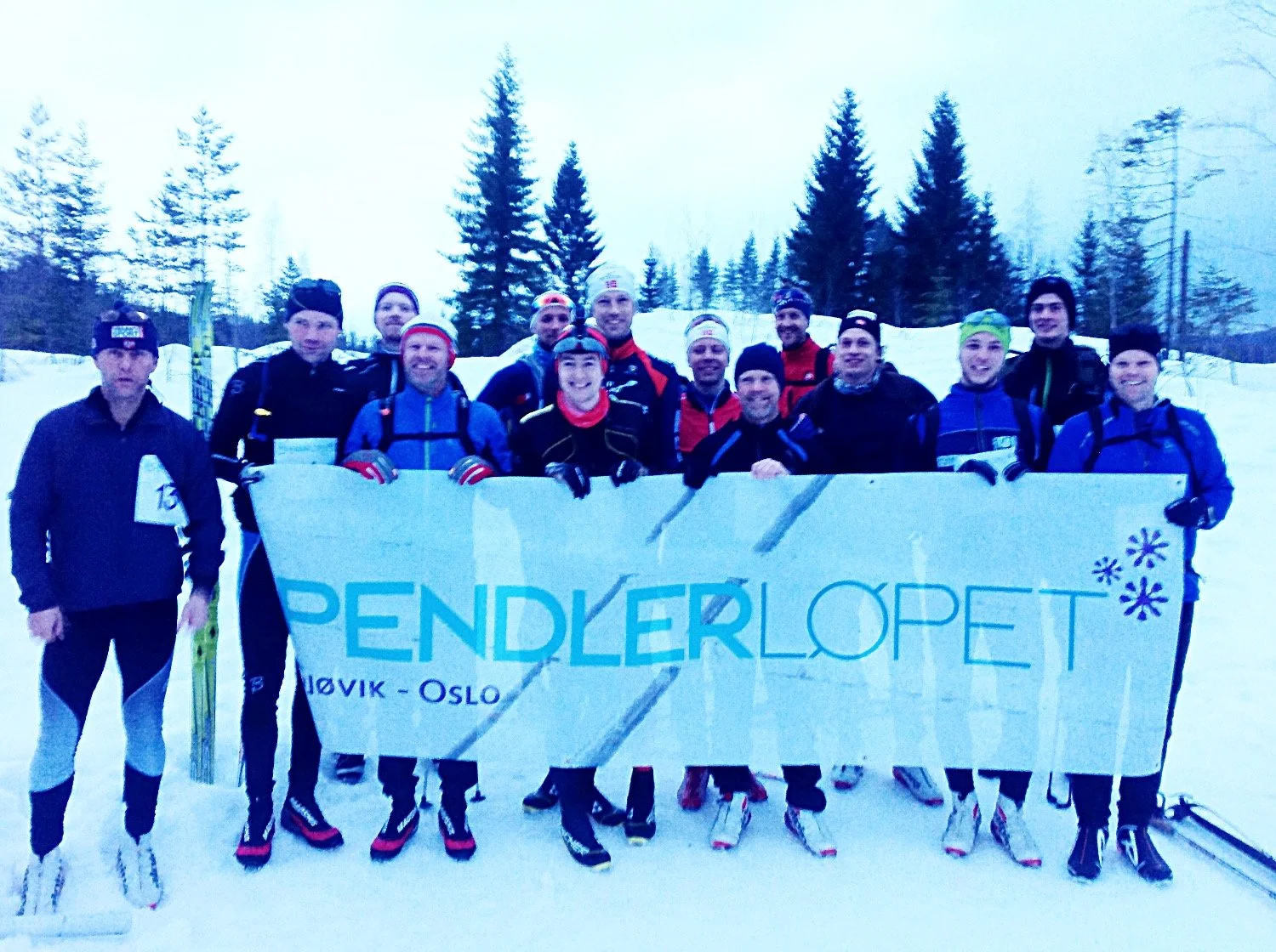 Group of people in winter clothing holding a banner with the text 'PENDLERLOPET' and standing on snow, with snowy landscape and evergreen trees in the background.