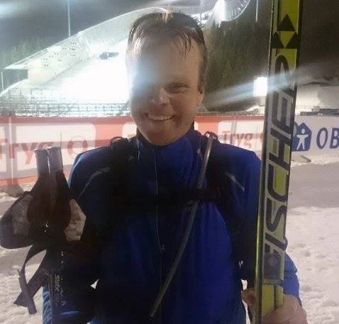 A smiling young man at an outdoor ski resort, holding skis and wearing a blue jacket.