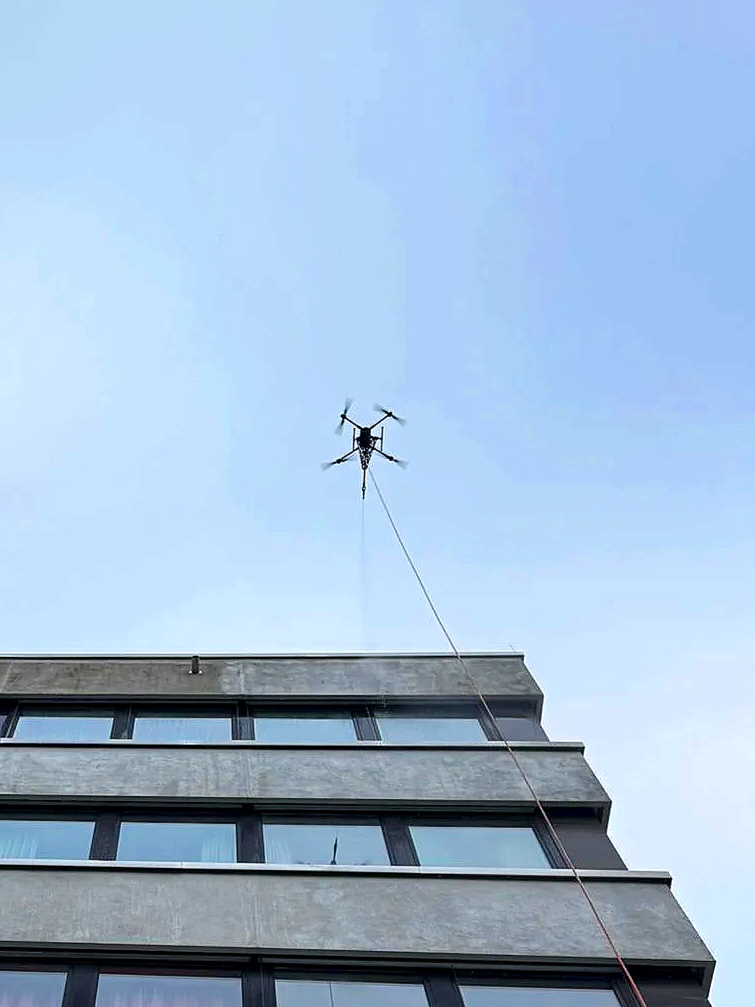 Drohne mit Seil hängt über einem mehrstöckigen Gebäude, blauer Himmel im Hintergrund.
