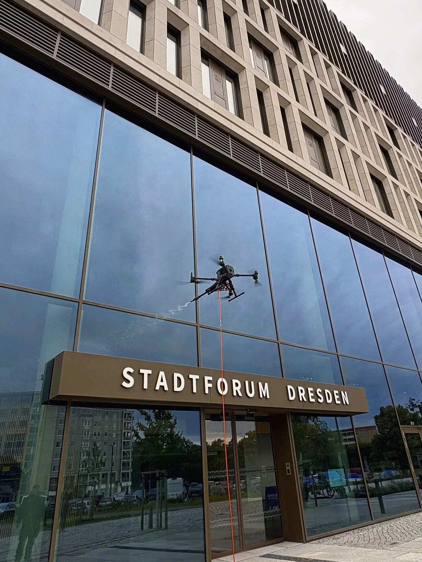 Ein Fensterputzer arbeitet an der Außenfassade des Stadtforums Dresden, während eine Drohne das Gebäude reinigt.