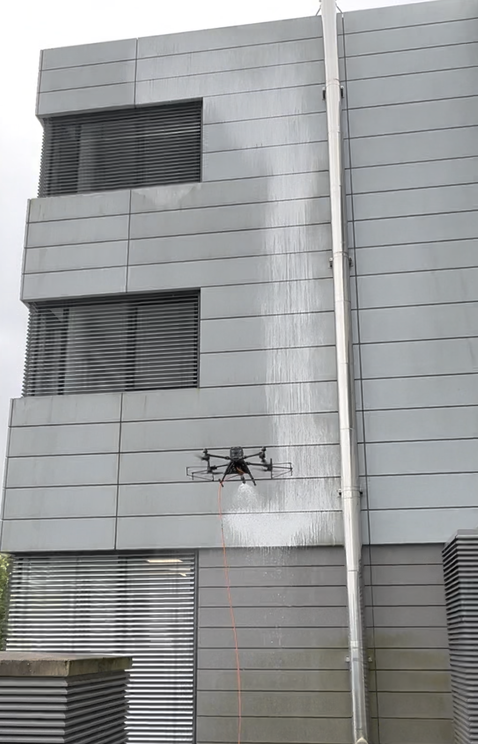 Ein Drohne reinigt die Außenfassade eines modernen, grauen Gebäudes mit horizontalen Linien.
