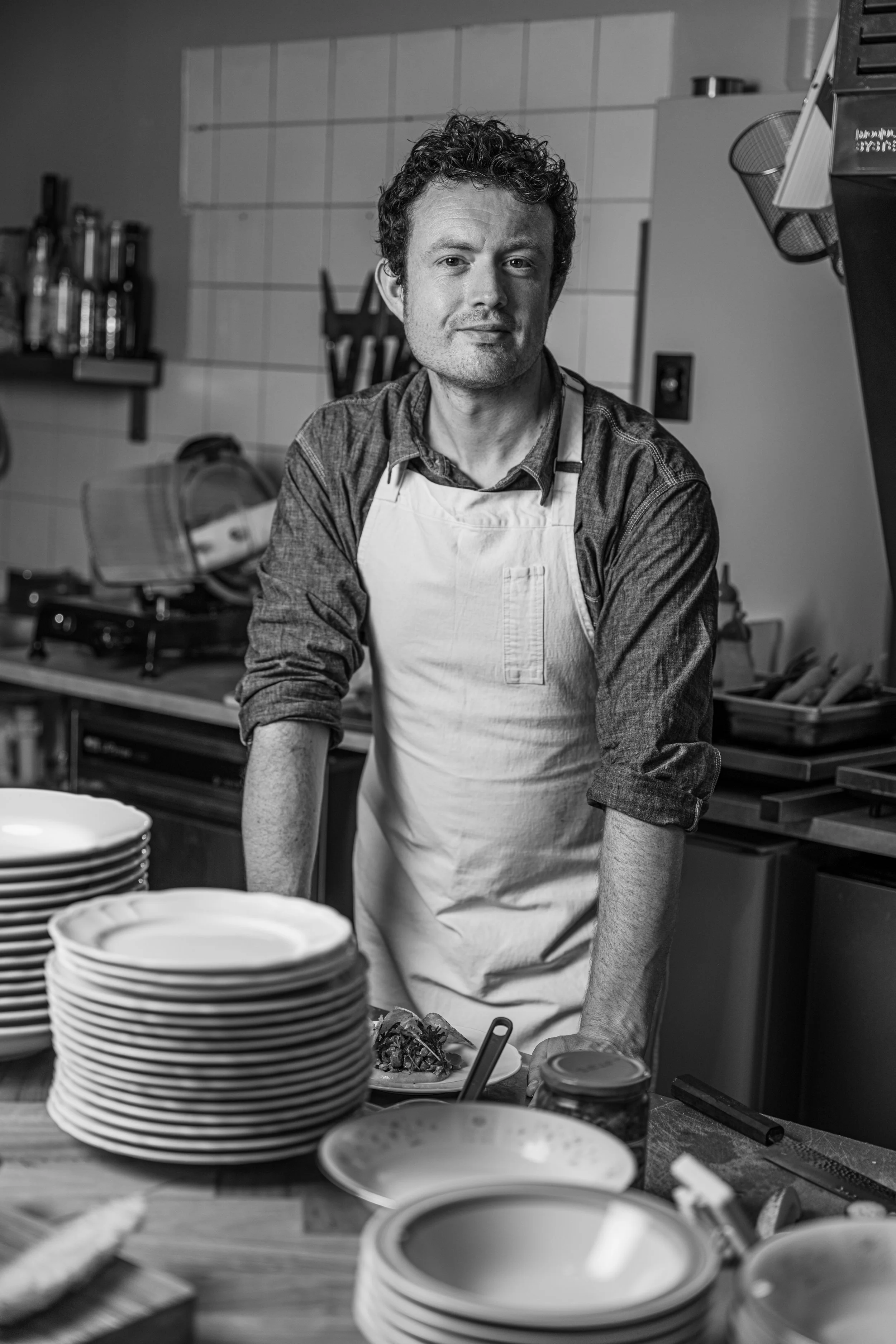 A man in a cooking apron standing in a kitchen with stacks of plates and various kitchen tools around him.