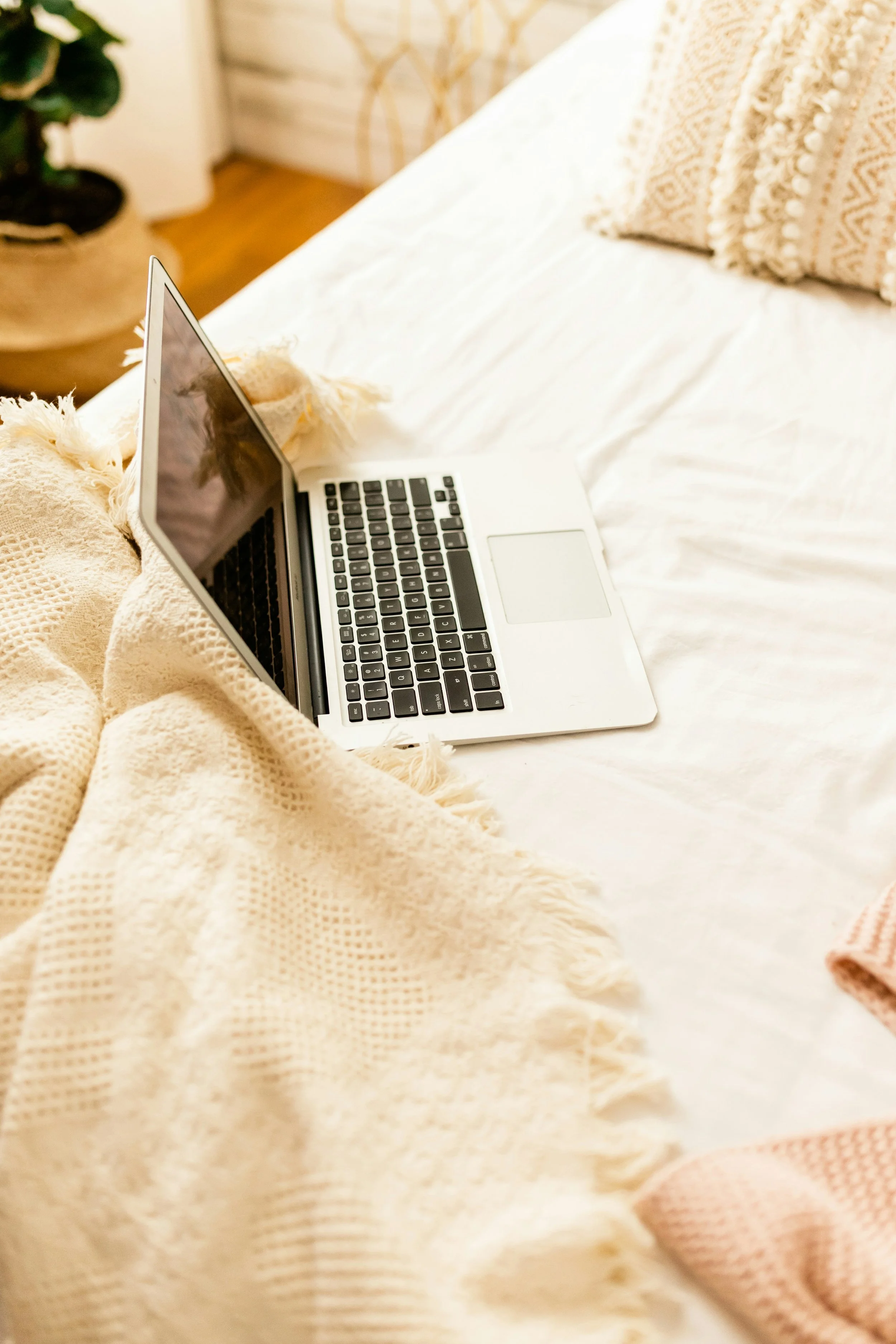 A silver laptop with black keyboard on a bed with white sheets and a beige knit blanket, with a decorative beige pillow nearby and a wooden nightstand with a plant in the background.