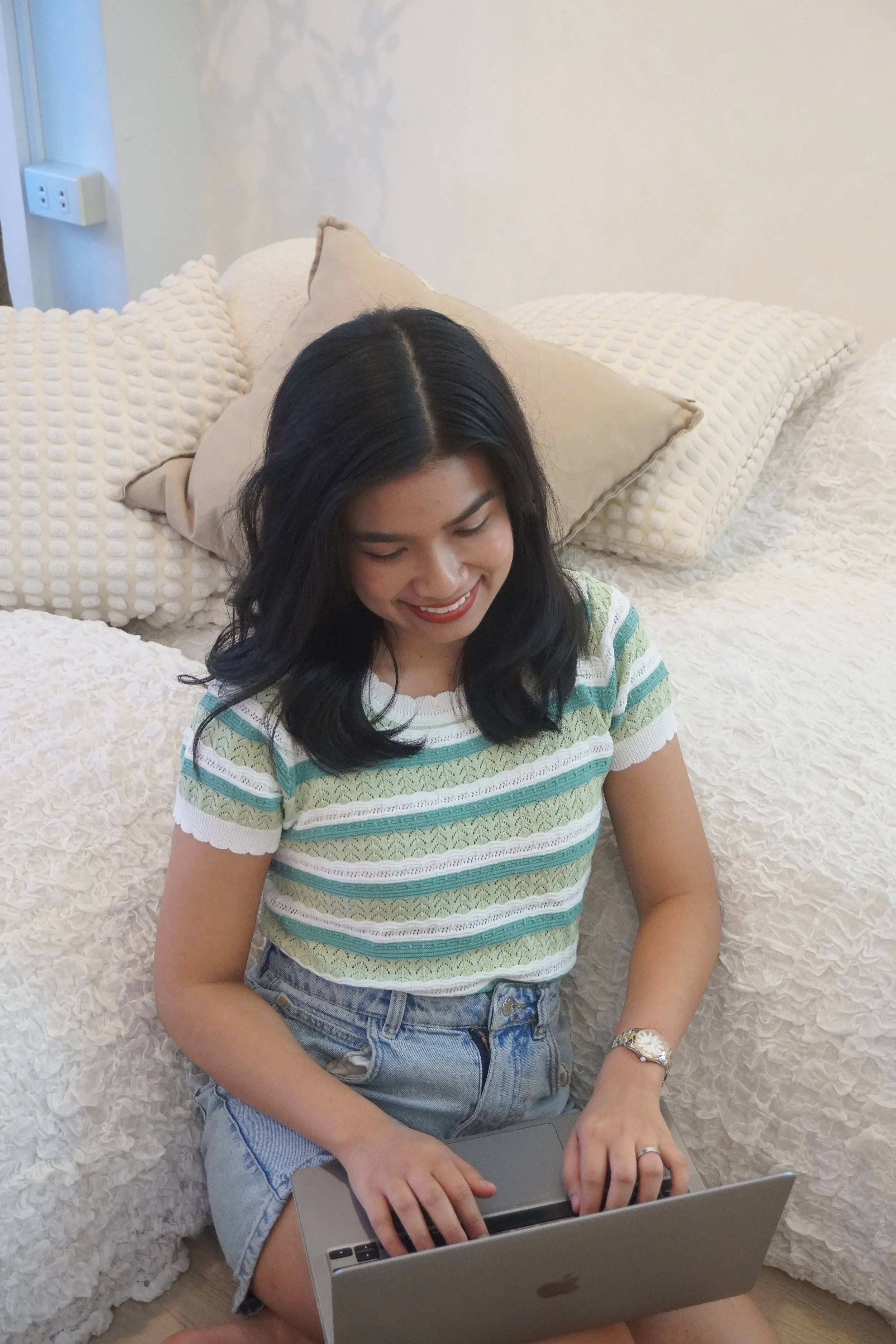 A woman sitting on the floor in front of a bed, using a laptop, smiling, with decorative pillows behind her.