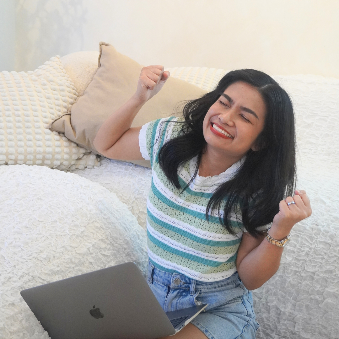 Happy woman sitting on bed with a laptop, raising her fists in celebration.