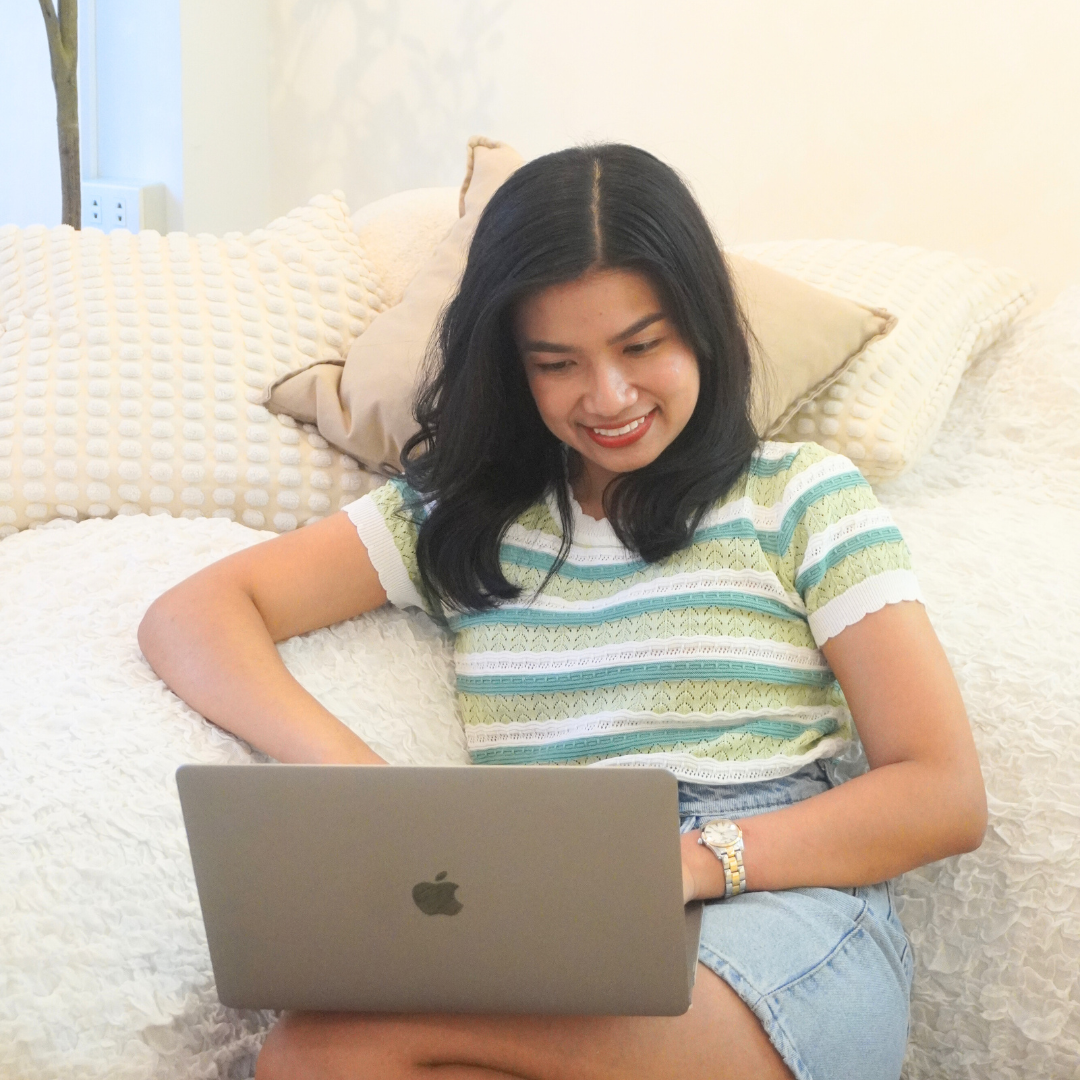 A young woman with black hair, wearing a striped green and white shirt and denim shorts, sitting on a bed with cream-colored bedding, smiling while looking at a silver MacBook laptop.