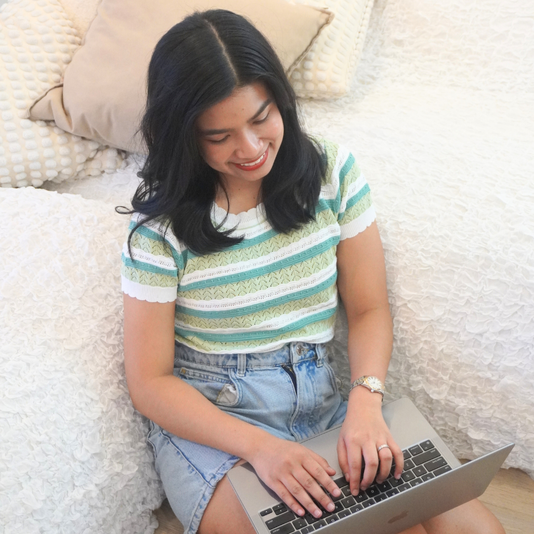 A woman sitting on a bed with white textured bedding, using a laptop, with pillows in the background.