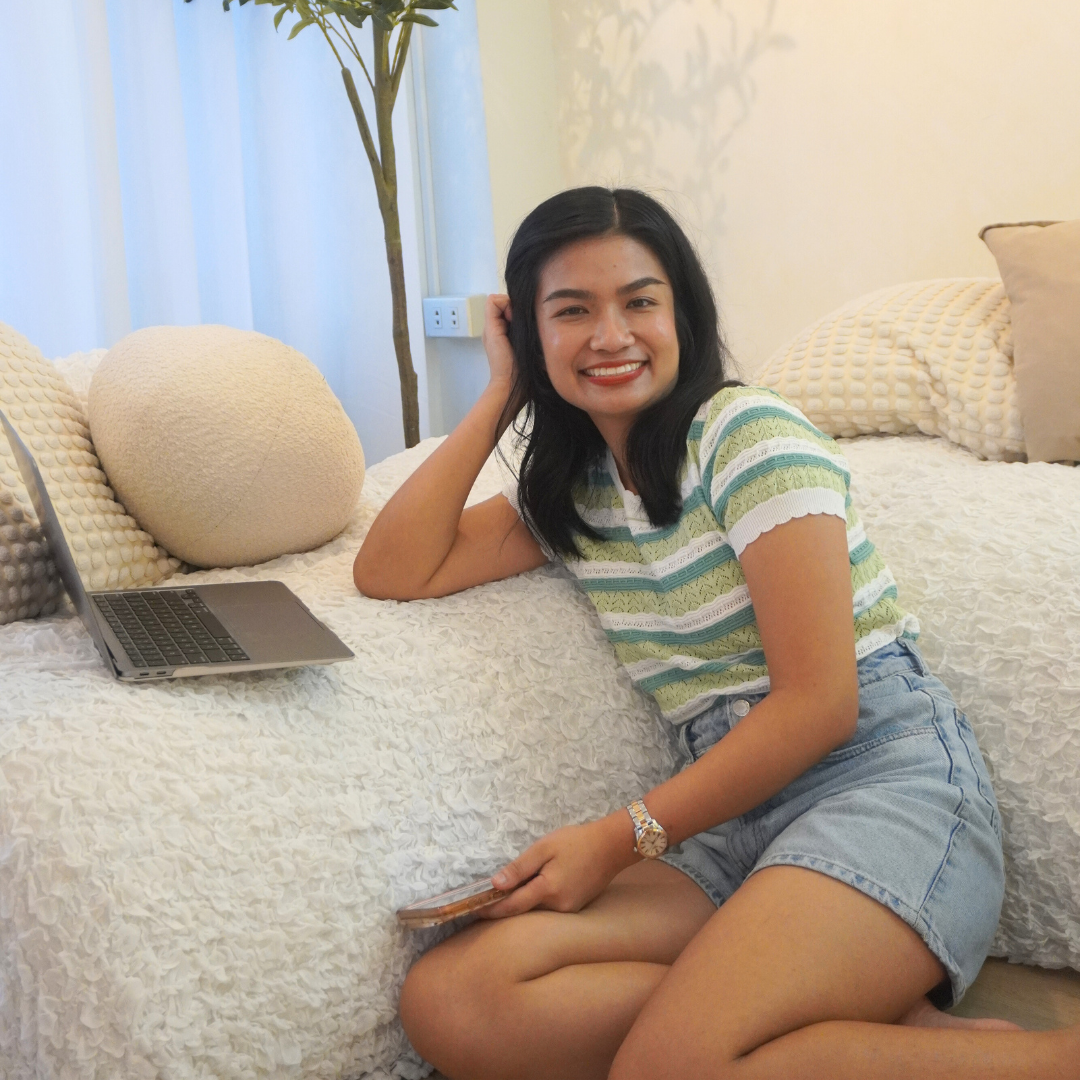 A young woman with dark hair smiling and lying on her side on a white textured bed in a well-lit room, holding a smartphone, with a laptop on the bed, beige pillows, a tall green plant, and a cream-colored wall in the background.