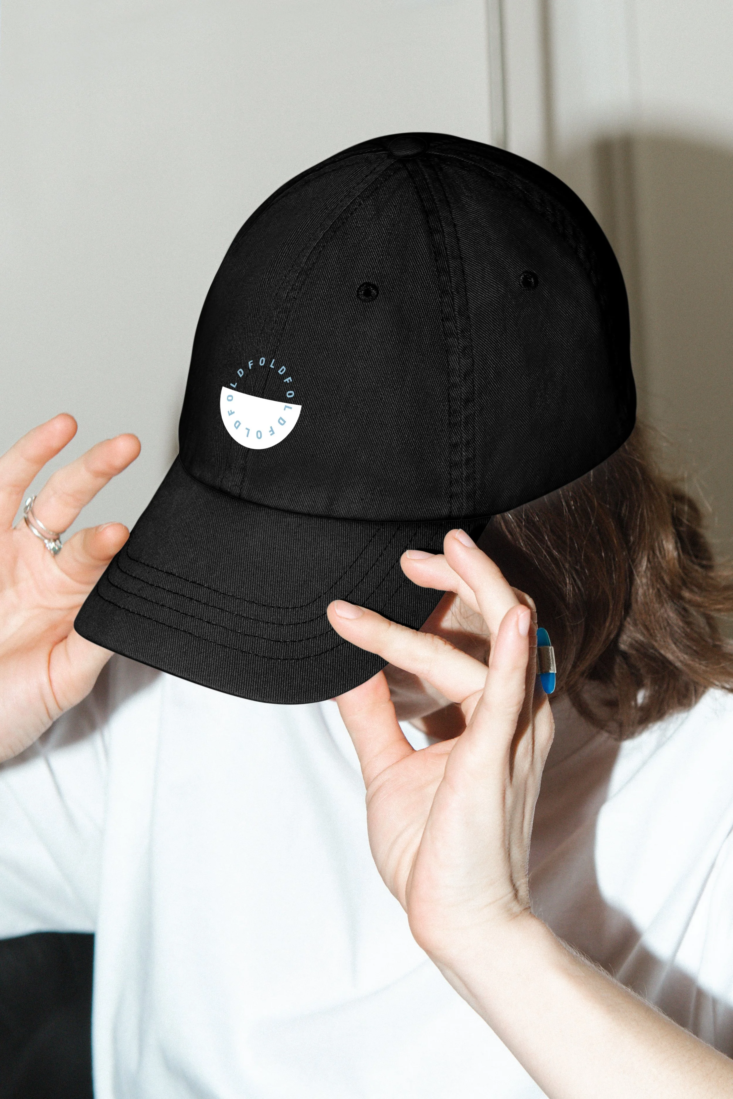 Person trägt eine schwarze Baseballkappe mit einem Logo. Die Person hält die Kappe vor das Gesicht, versteckt es teilweise. Die Person trägt eine weiße Kleidung und hat braune, lockige Haare