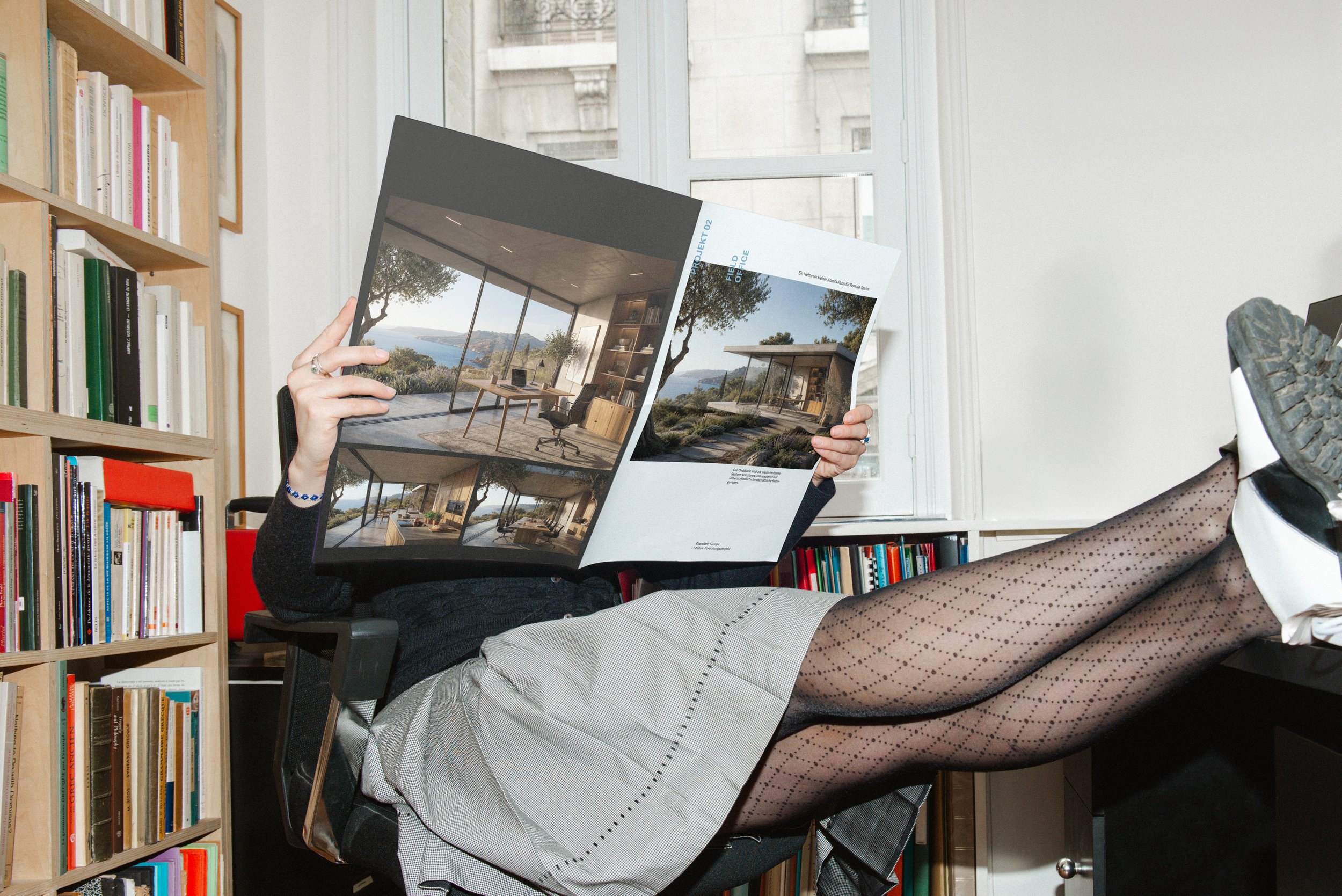 Eine Person liegt in einem Bürostuhl mit hochgelegten Beinen und liest eine Broschüre mit Bildern von modernen Häusern. Im Hintergrund befindet sich ein Bücherregal und ein Fenster, durch das ein Gebäude zu sehen ist.