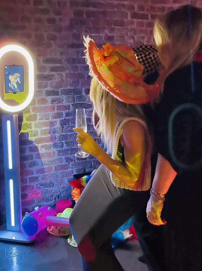 A woman with a large unicorn-patterned sombrero hat is sitting on a chair, holding a champagne flute, in front of a brick wall with colorful party decorations and balloons on the floor.