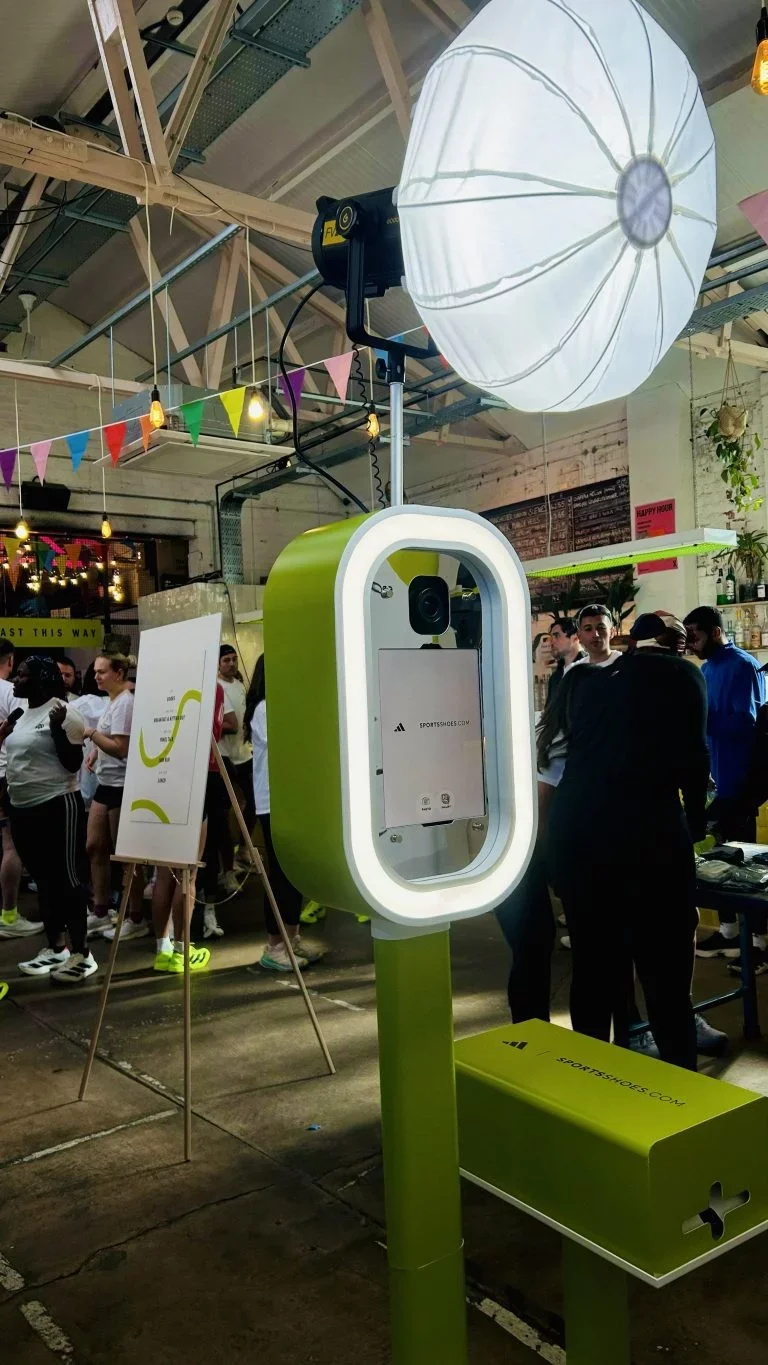 Photo of a photo booth with a large light inside a warehouse-like venue decorated with colorful bunting and lights, with people gathered around in the background.