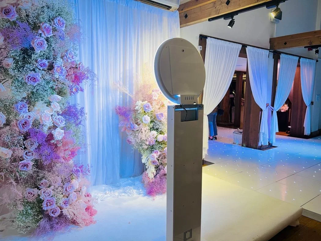 Wedding or event backdrop with pink and purple roses and white drapery, photo booth in the foreground, decorated venue with white curtains and string lights.