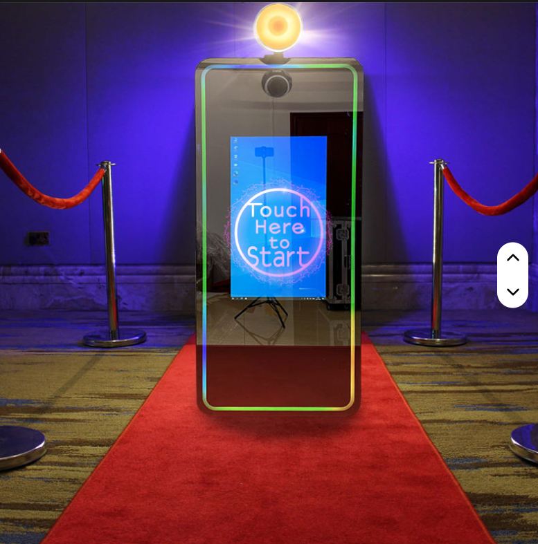 A mirror-like kiosk with a digital screen displaying 'Touch Here to Start' surrounded by a neon circle, placed on a red carpet with velvet ropes in a dimly lit room.