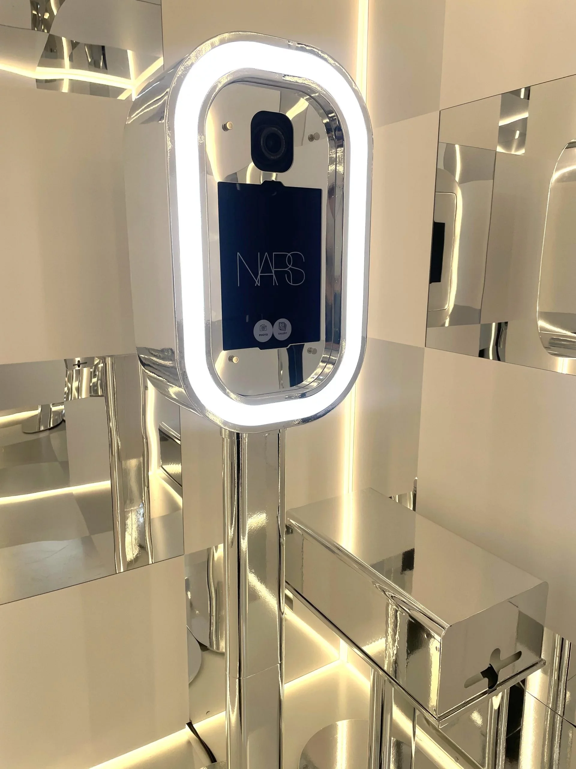 A high-tech, futuristic photo booth or kiosk with a sleek, silver, and reflective exterior, featuring a large vertical screen labeled 'NARS' and a camera above it, situated in a modern, well-lit environment.