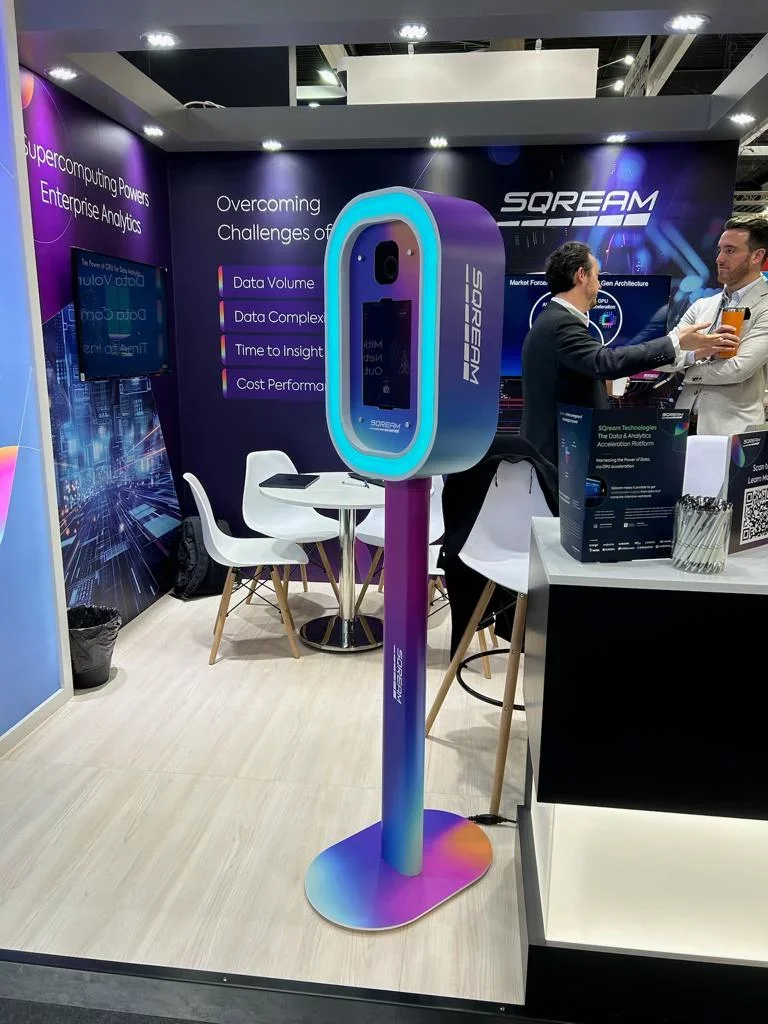 A booth at a technology exhibition demonstrating a high-tech device called SOREAM. The device appears to be a large, futuristic, vertical screen or scanner with blue and purple lighting effects. There are two men engaging in conversation to the right of the device, and the background includes promotional banners highlighting data analytics and computing solutions.