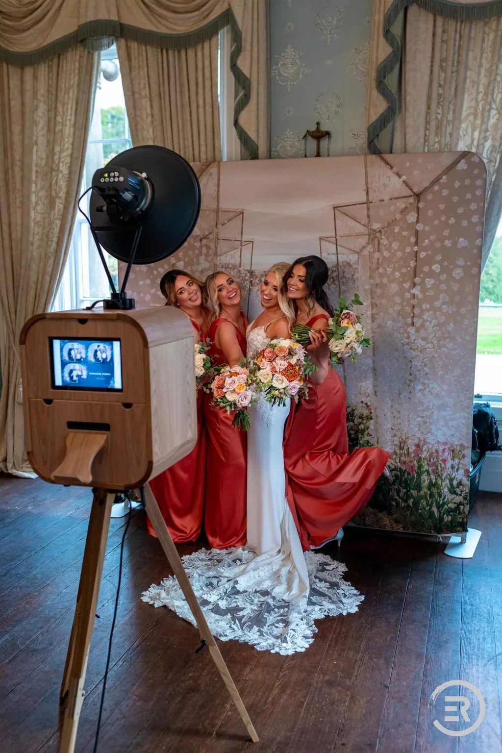 Four women at a wedding, three bridesmaids in matching red dresses and a bride in a white dress, holding bouquets of flowers, smiling for a photo.