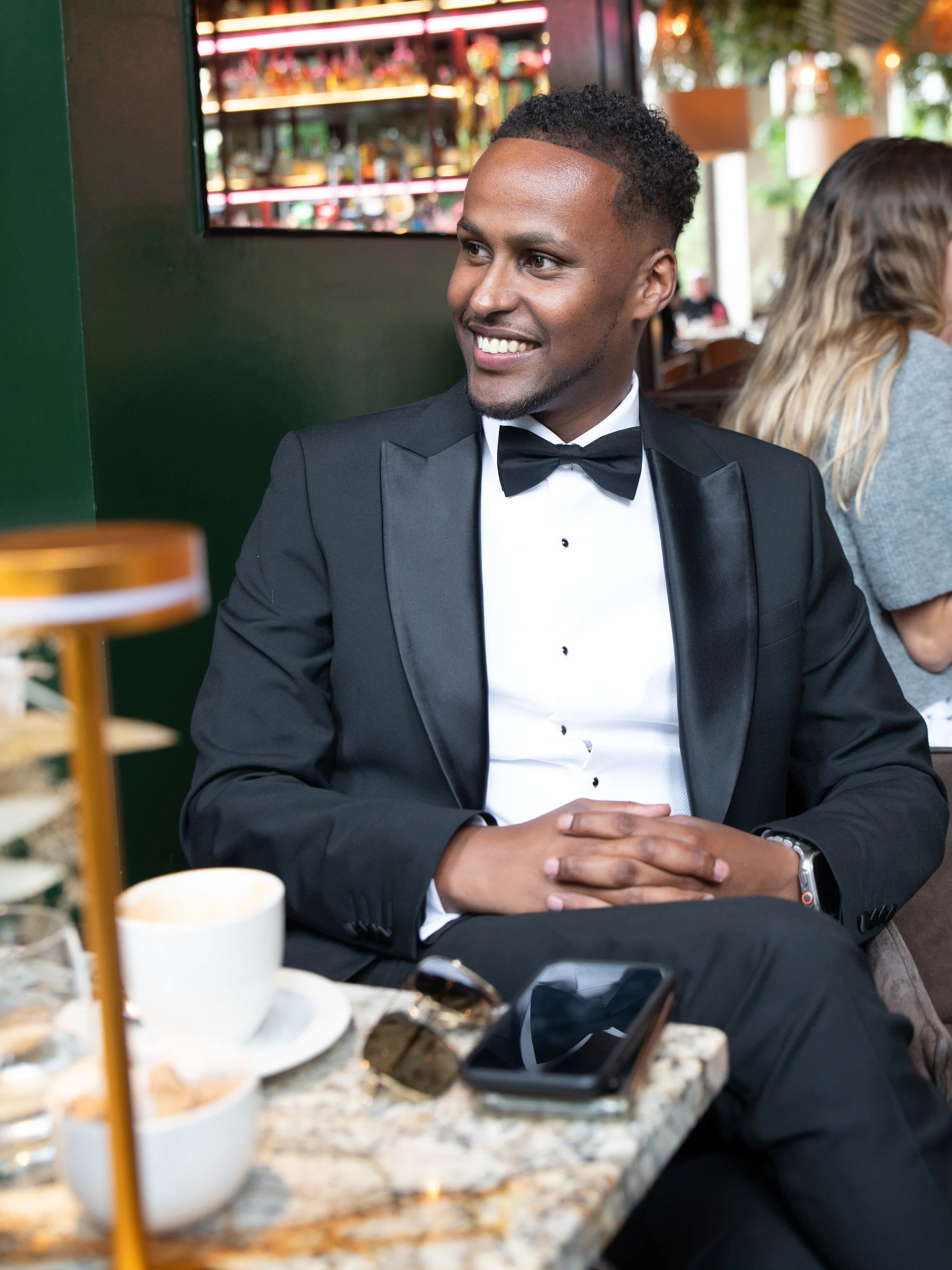 A man in a black tuxedo with a bow tie, smiling, sitting at a table in a restaurant.