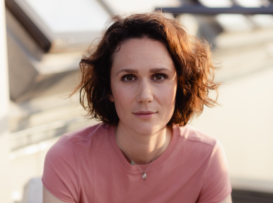 Porträt einer jungen Frau mit kurzen, lockigen braunen Haaren, in einem rosa T-Shirt, mit Sonnenbrillen und einem Wasserhahn im Hintergrund.