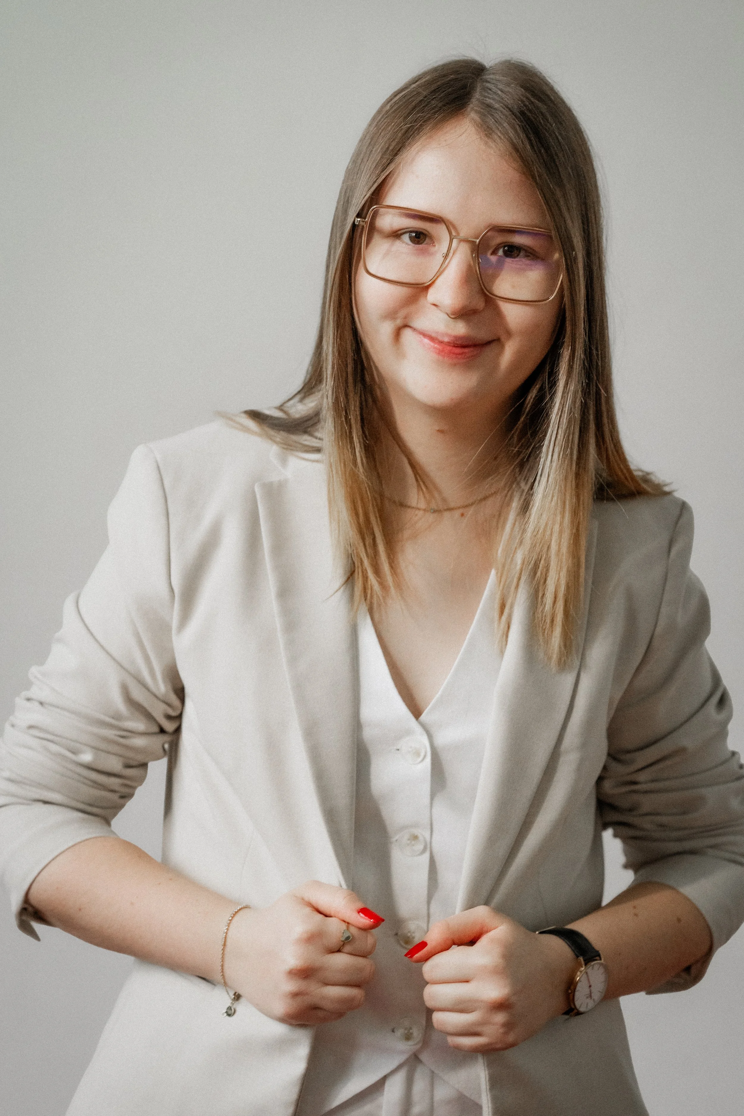 Junge Frau mit Brille, in beige Blazer und weißem Hemd, lächelnd, vor neutralem Hintergrund.