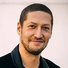 Ein Mann mit dunklem, kurz geschnittenem Haar und Bart, trägt ein dunkles Hemd und lächelt leicht.